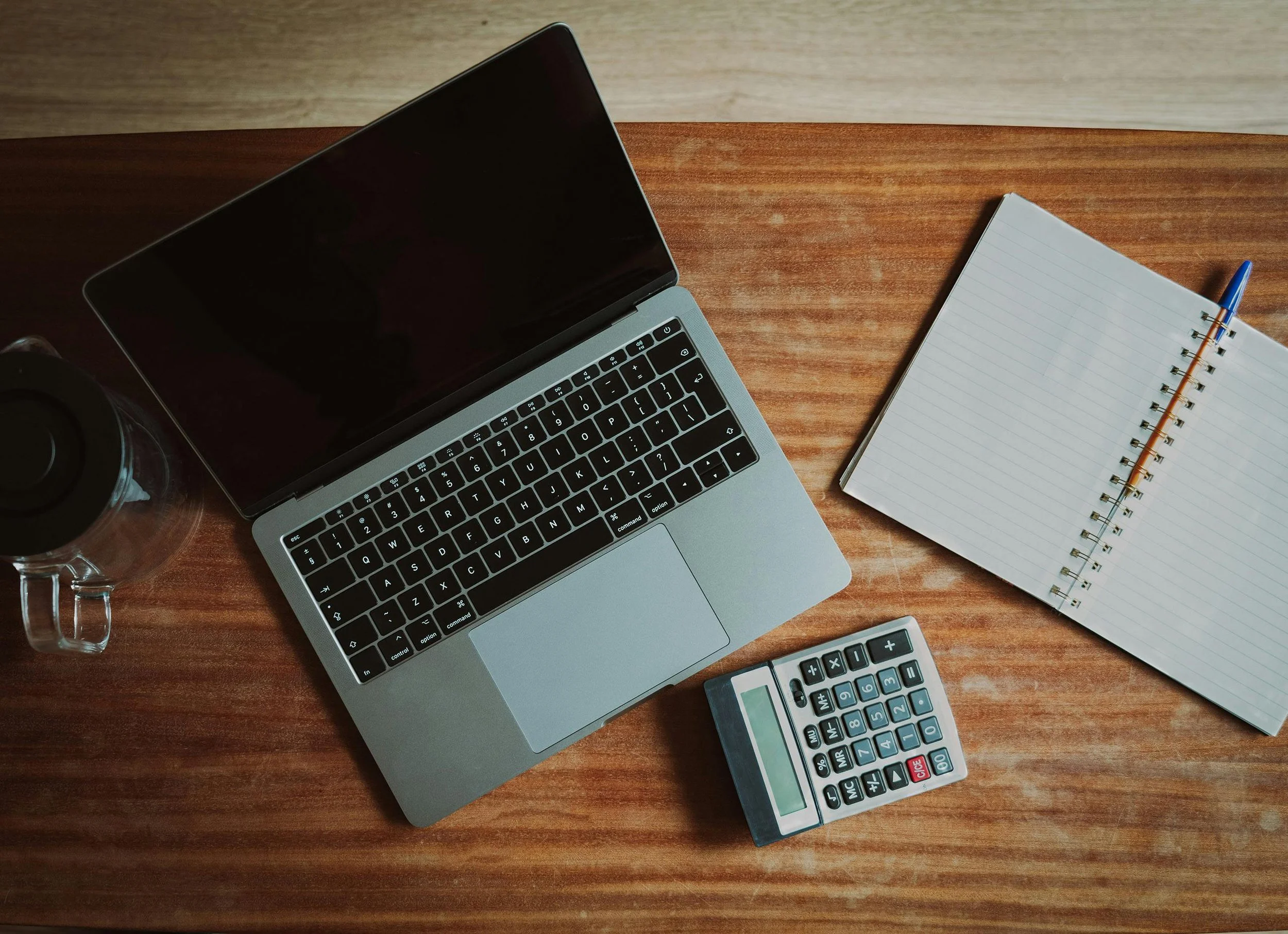 A laptop, a calculator, a spiral notebook with lined pages, a blue pen, and a glass of water on a wooden desk. a typical accounting setup
