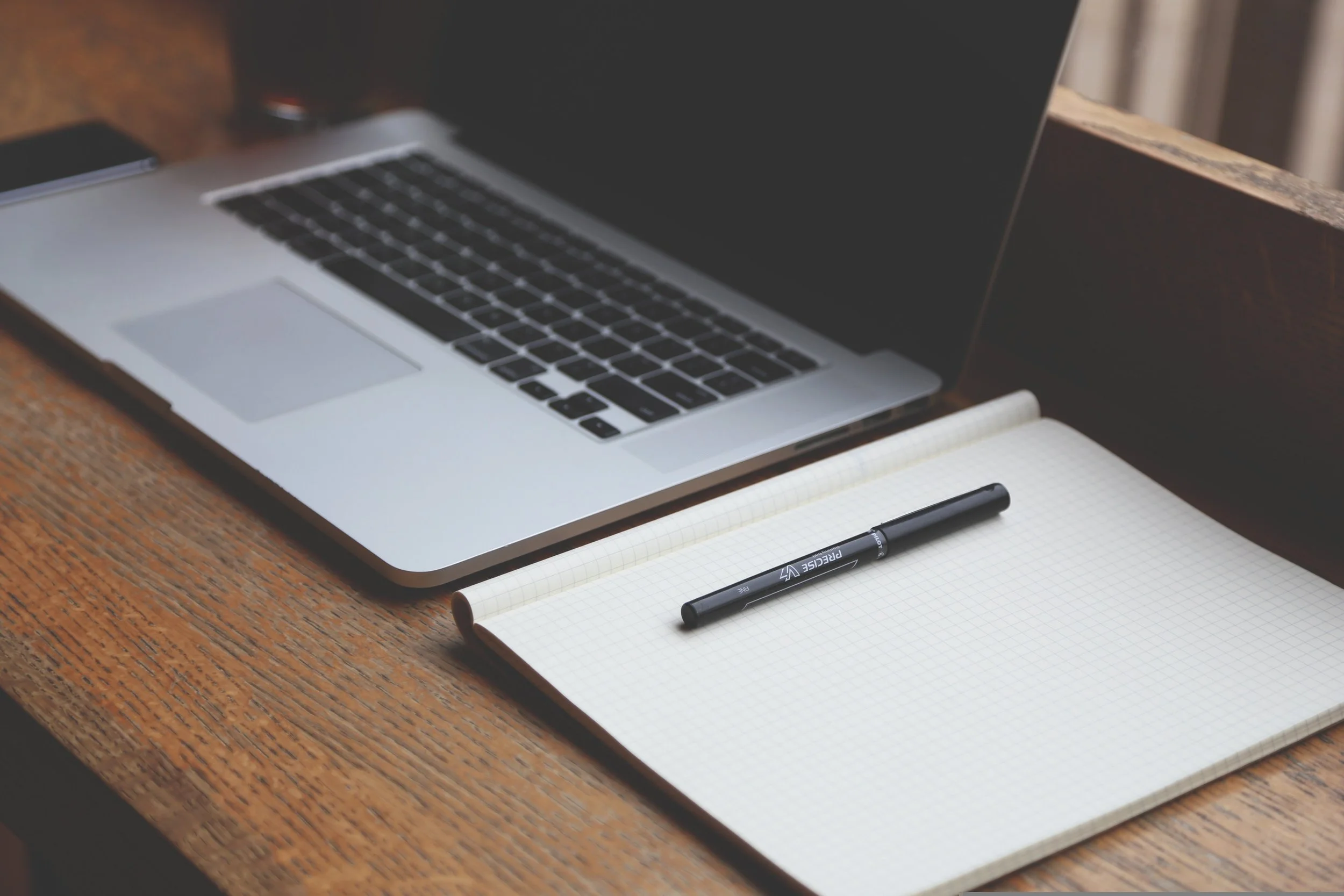 Open silver laptop with black keyboard next to a black pen on a white grid notebook, on a wooden desk.