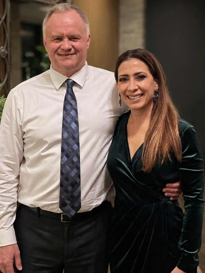 A man and a woman standing together, smiling at the camera indoors. The man is wearing a white shirt and plaid tie, and the woman is in a black velvet dress with long hair and earrings.