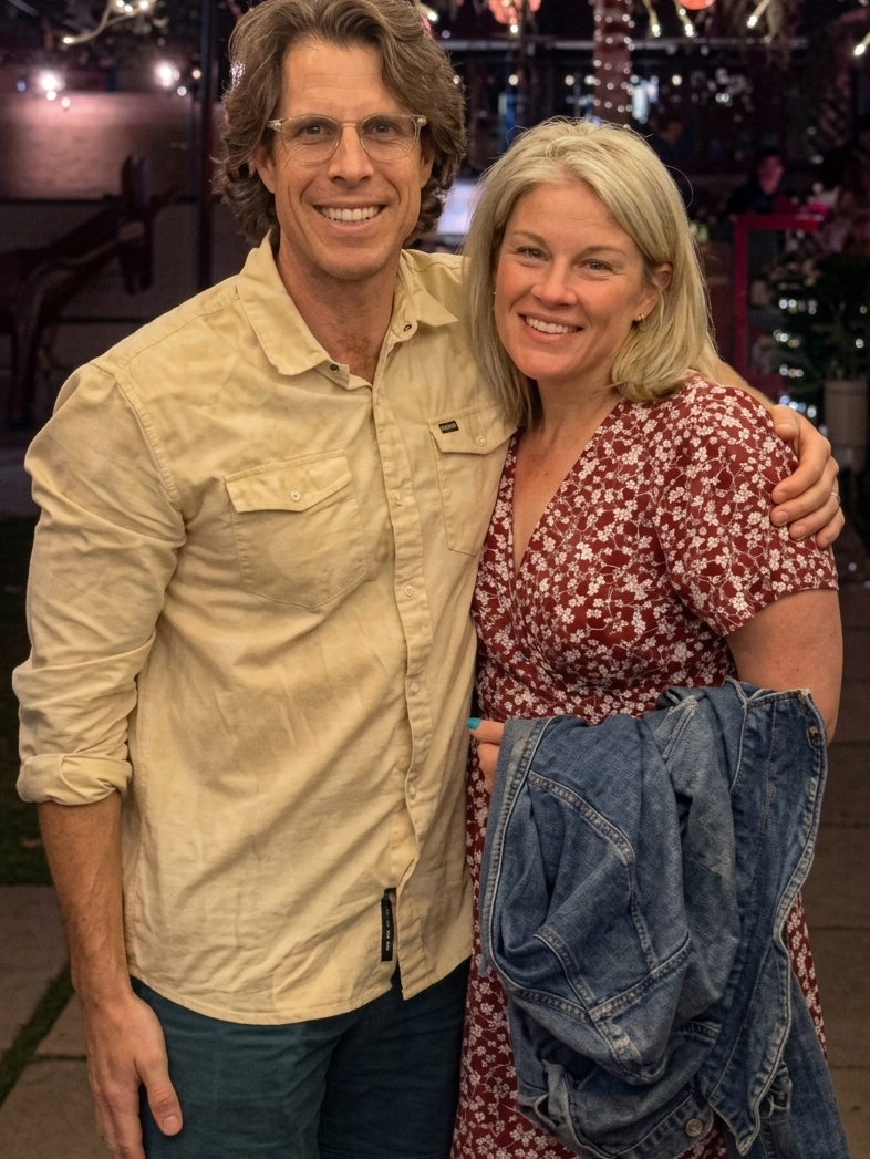 A smiling man and woman standing close together at night, with the man’s arm around the woman’s shoulder. The man has wavy brown hair, glasses, and is wearing a beige button-up shirt. The woman has shoulder-length blonde hair, and is dressed in a red and white floral dress, holding a denim jacket in her hand. The background features blurred string lights and a decorated outdoor setting.