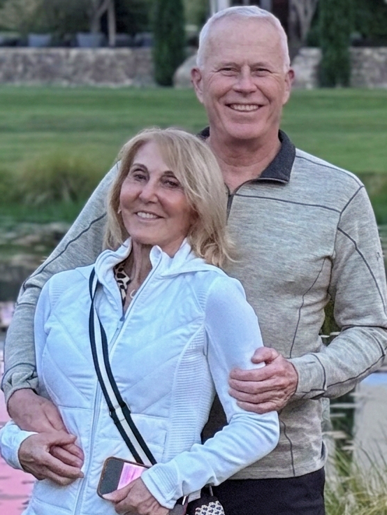 A smiling older man and woman standing outdoors near water, with the man hugging the woman from behind, both wearing casual athletic clothing.