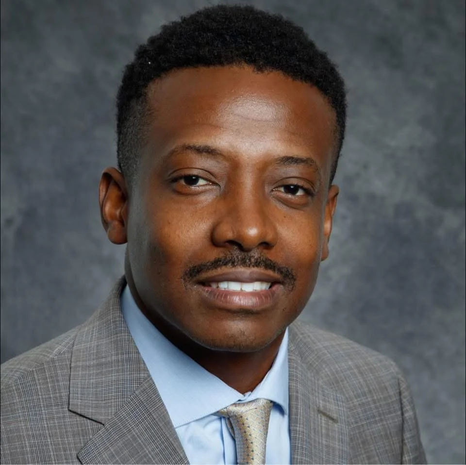 A man in a gray suit with a light blue shirt and a patterned tie, posing for a professional portrait against a dark, textured background.