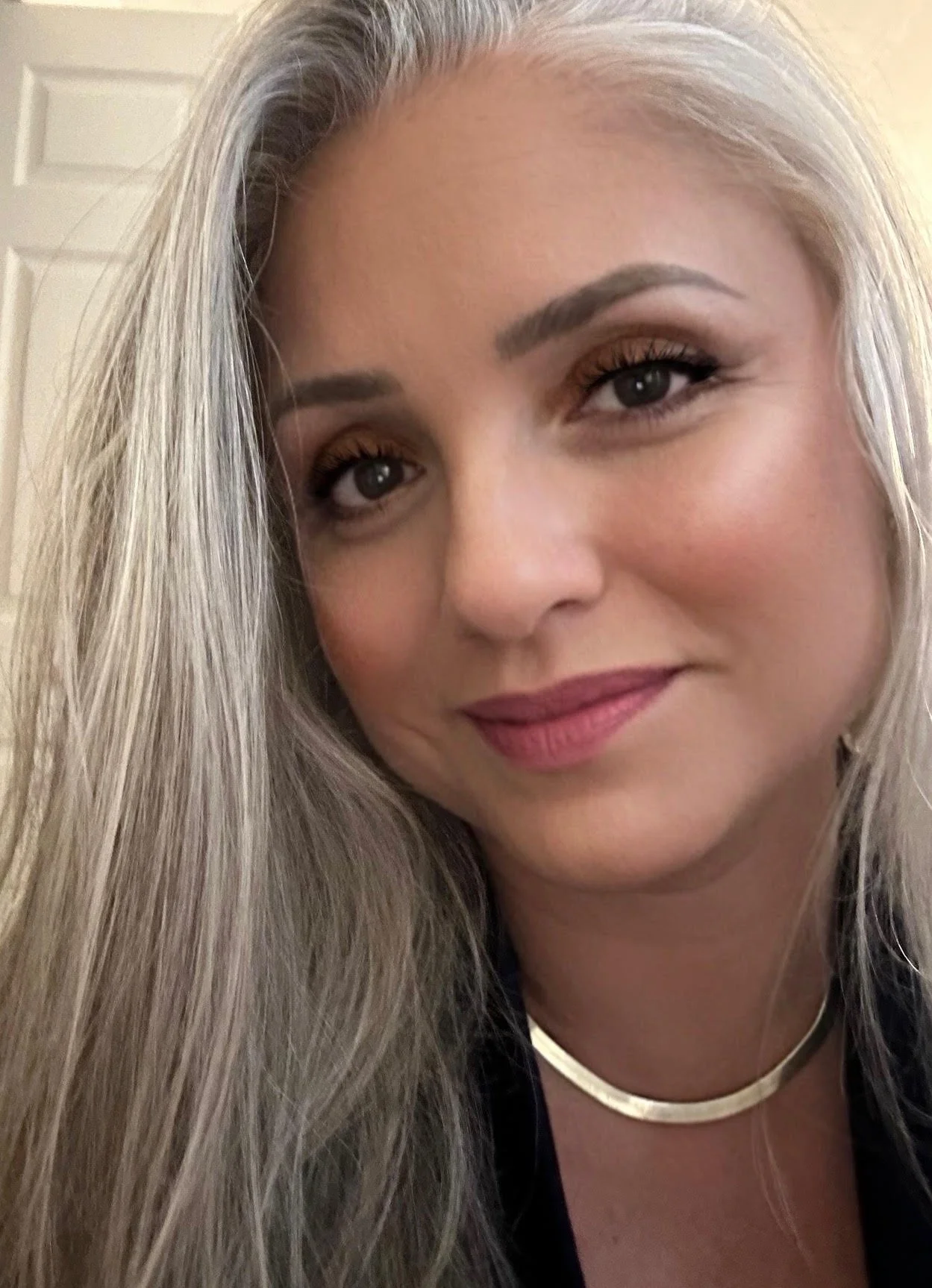 A close-up of a woman with long, blonde hair, smiling softly, wearing makeup, a gold choker necklace, and a dark top, indoors with a beige background.