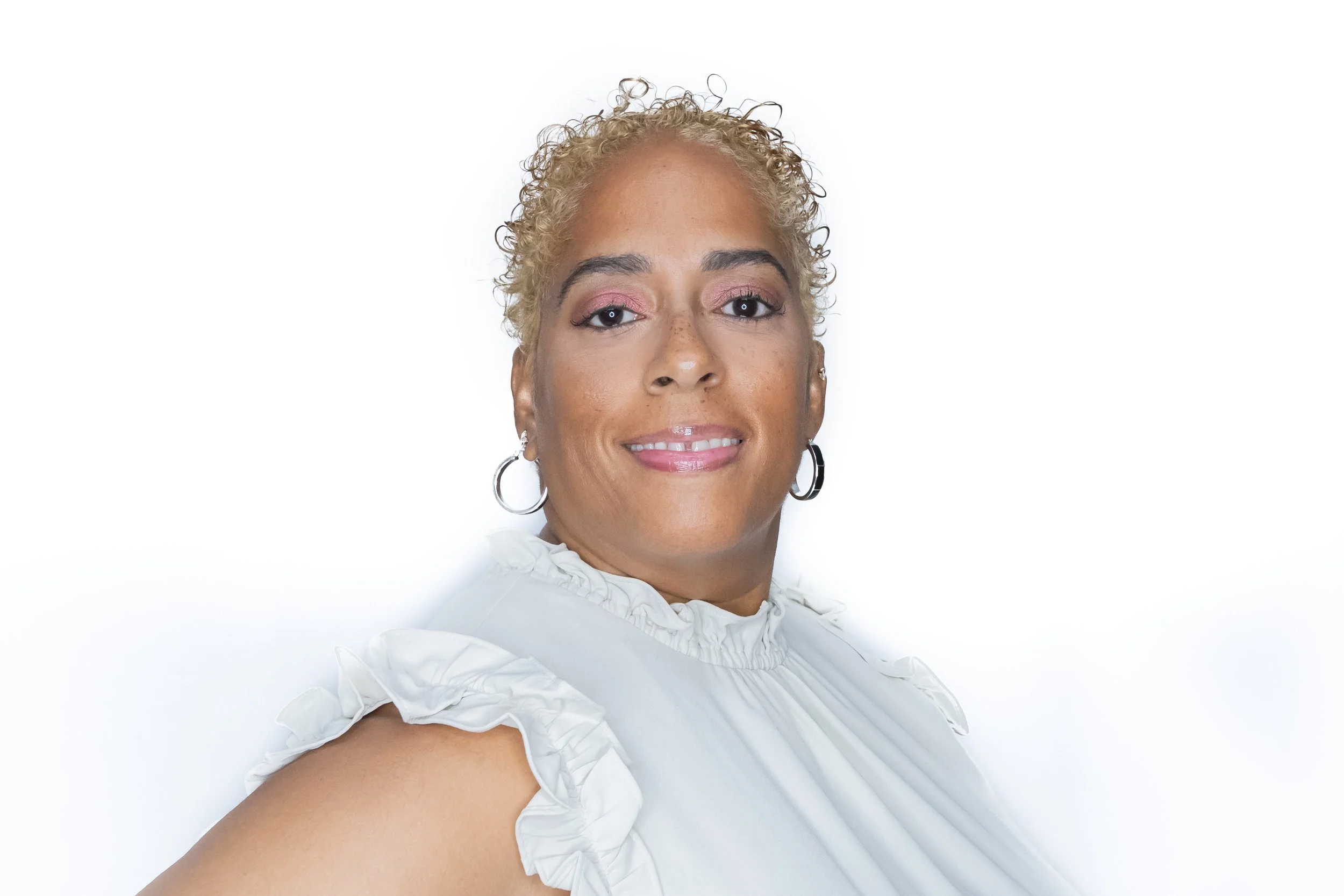 Portrait of a smiling woman with short, curly blonde hair, wearing hoop earrings and a white ruffled blouse, against a white background.