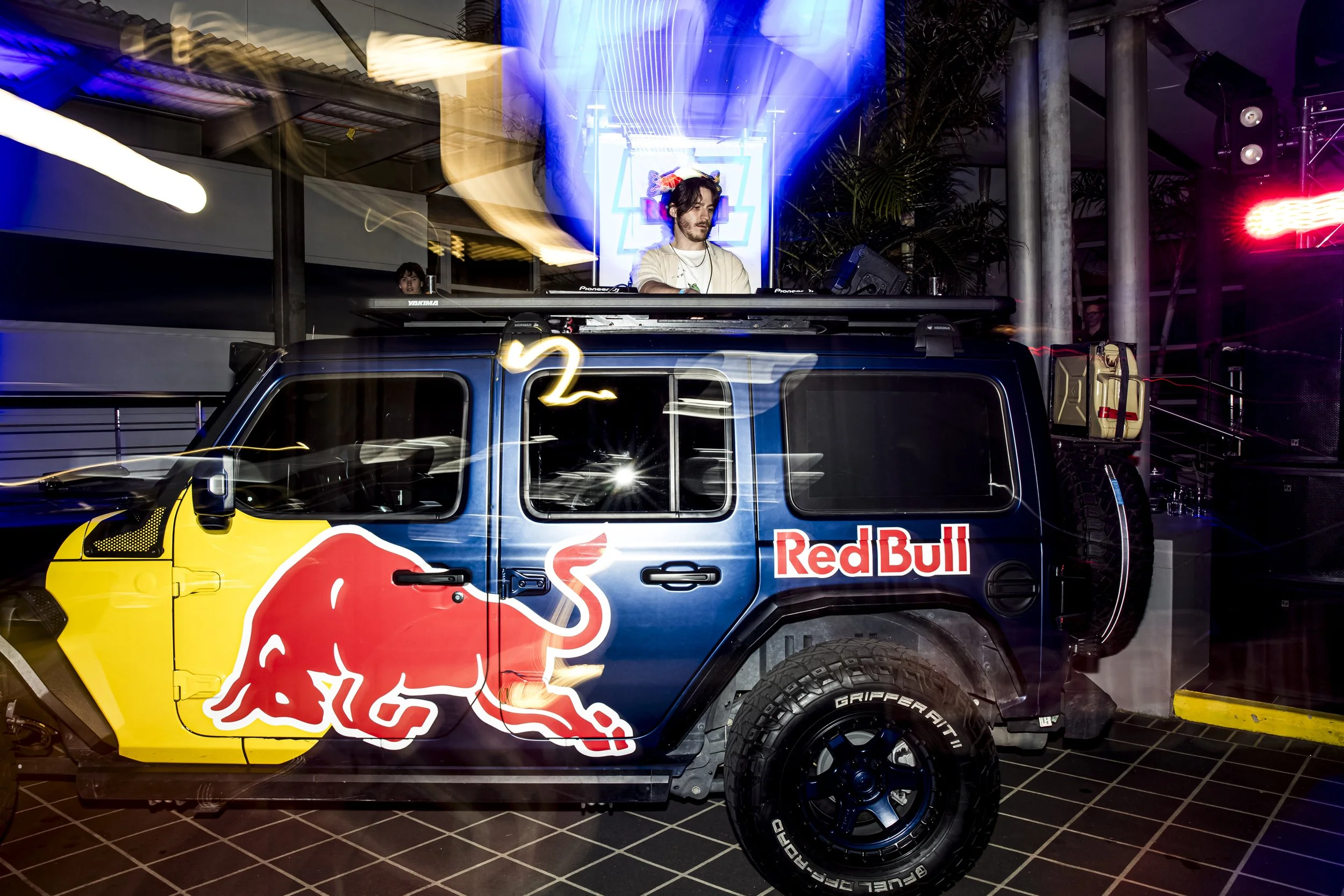 A person DJing on top of a Red Bull branded vehicle with a blue and yellow color scheme and a Red Bull logo, located indoors with neon lights and metallic structural elements.