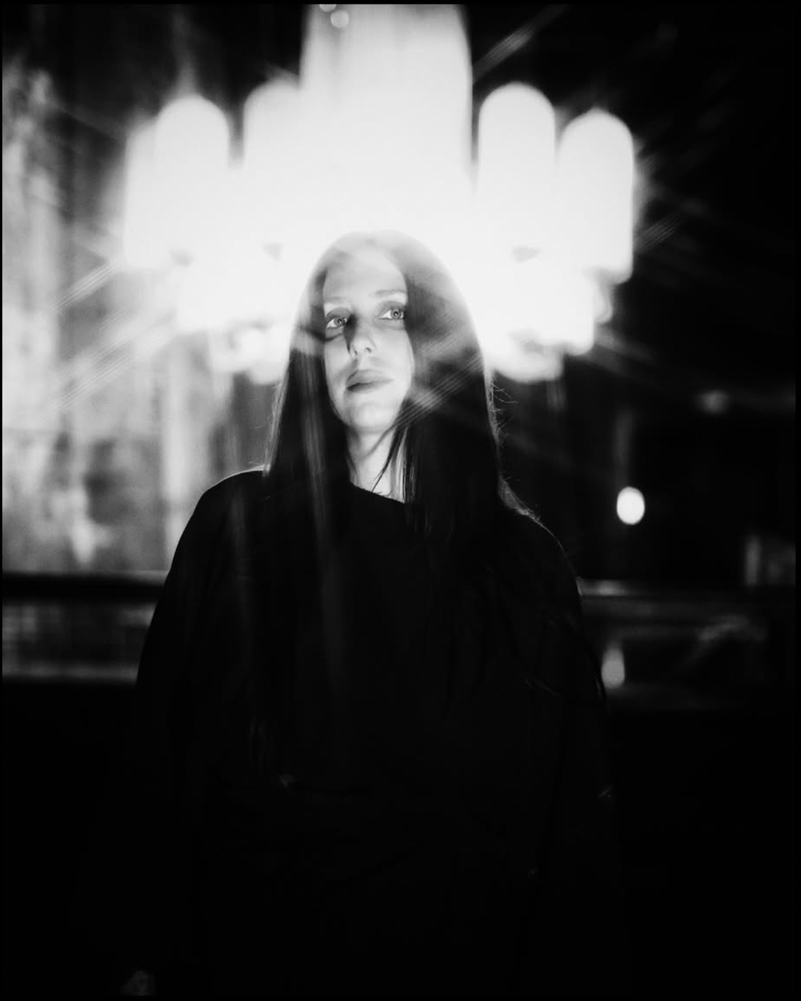 Black and white photo of a woman with long dark hair standing in front of a bright neon sign with three fingers showing. The woman's face is illuminated, with a serious expression, and the background is blurry.