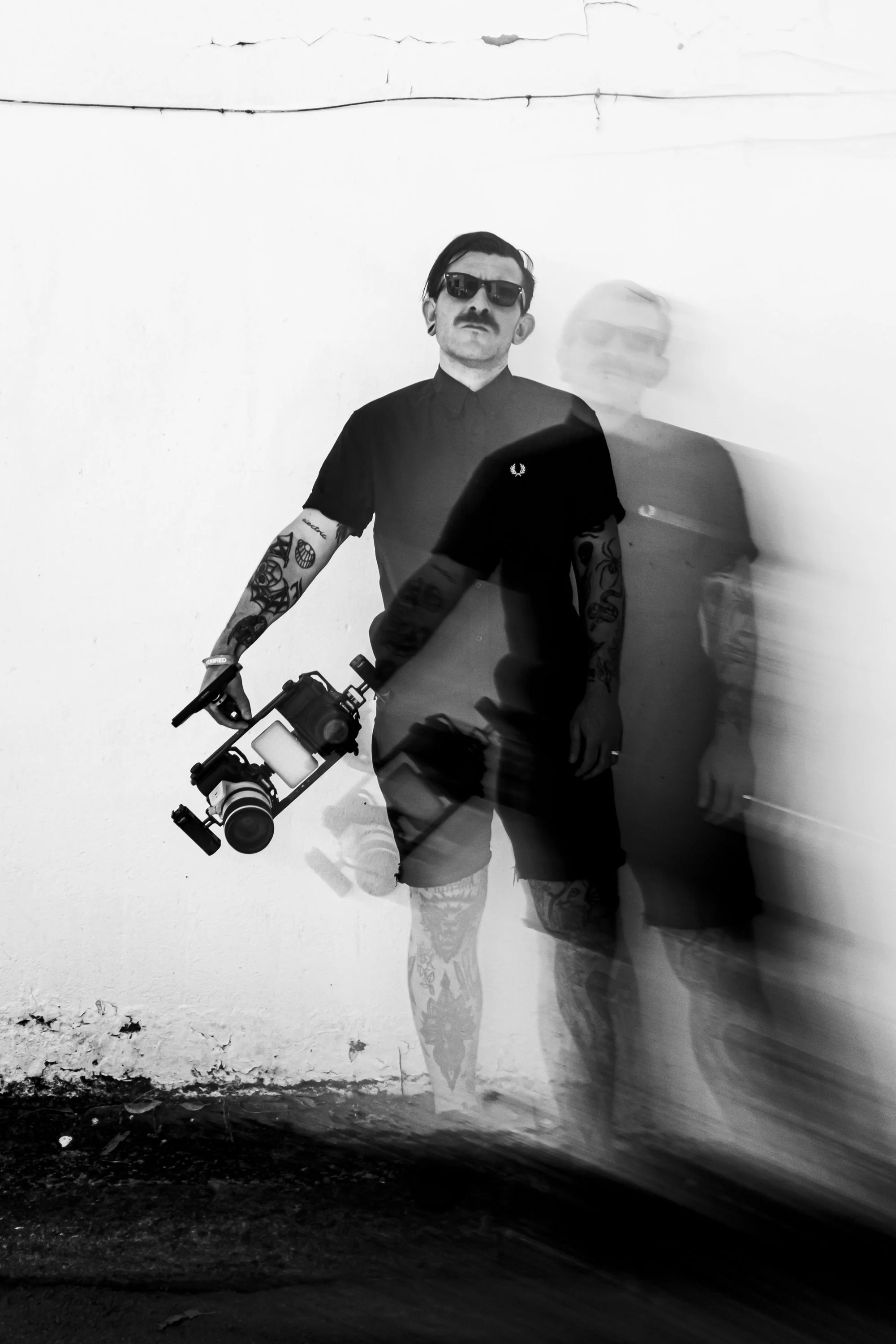 Black and white photo of a man with sunglasses, tattoos, and a mustache holding a camera on a tripod, standing against a plain wall with a reflection creating a ghostly duplicate.