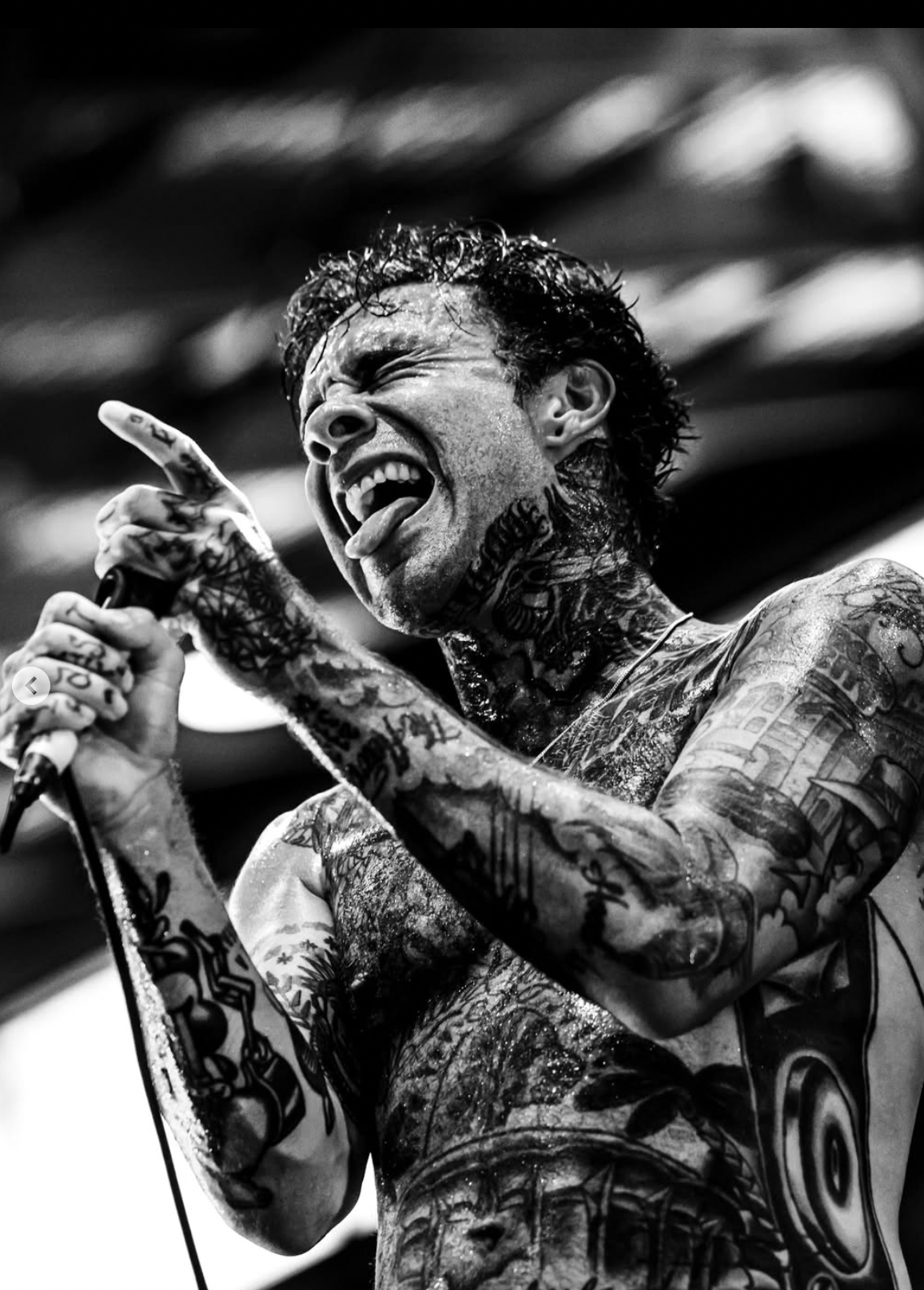 Black and white photo of a heavily tattooed male singer passionately singing into a microphone with his mouth open and tongue out, holding the microphone with one hand.