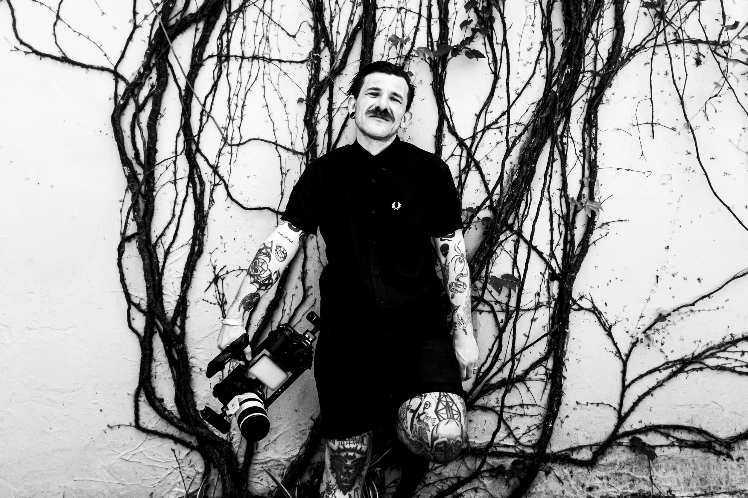 A man with tattoos on his arms and legs, holding a professional camera, standing in front of a wall with tangled vines, in black and white.