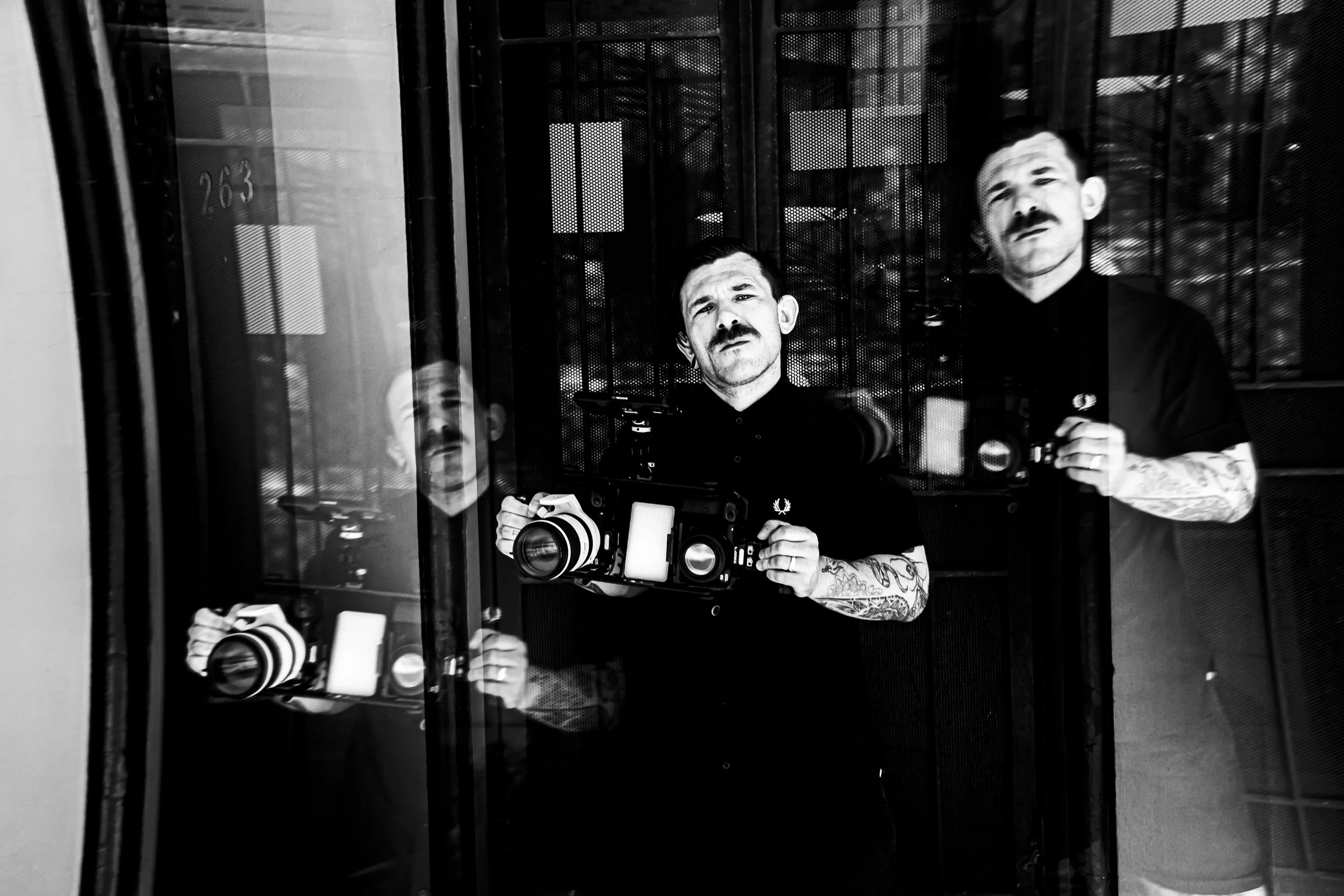 A reflection of a man with tattoos on his arms, holding a camera, in a glass window or door, with multiple reflections of himself and another man in the background.