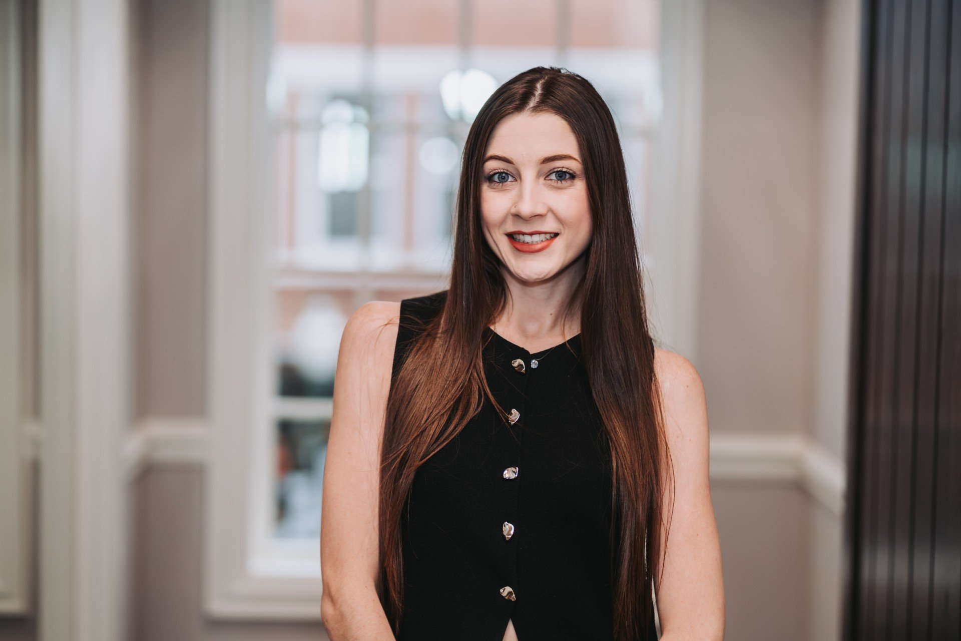 A young woman with long brown hair, wearing a black sleeveless top with button details, standing indoors in front of a window with blurred buildings outside.