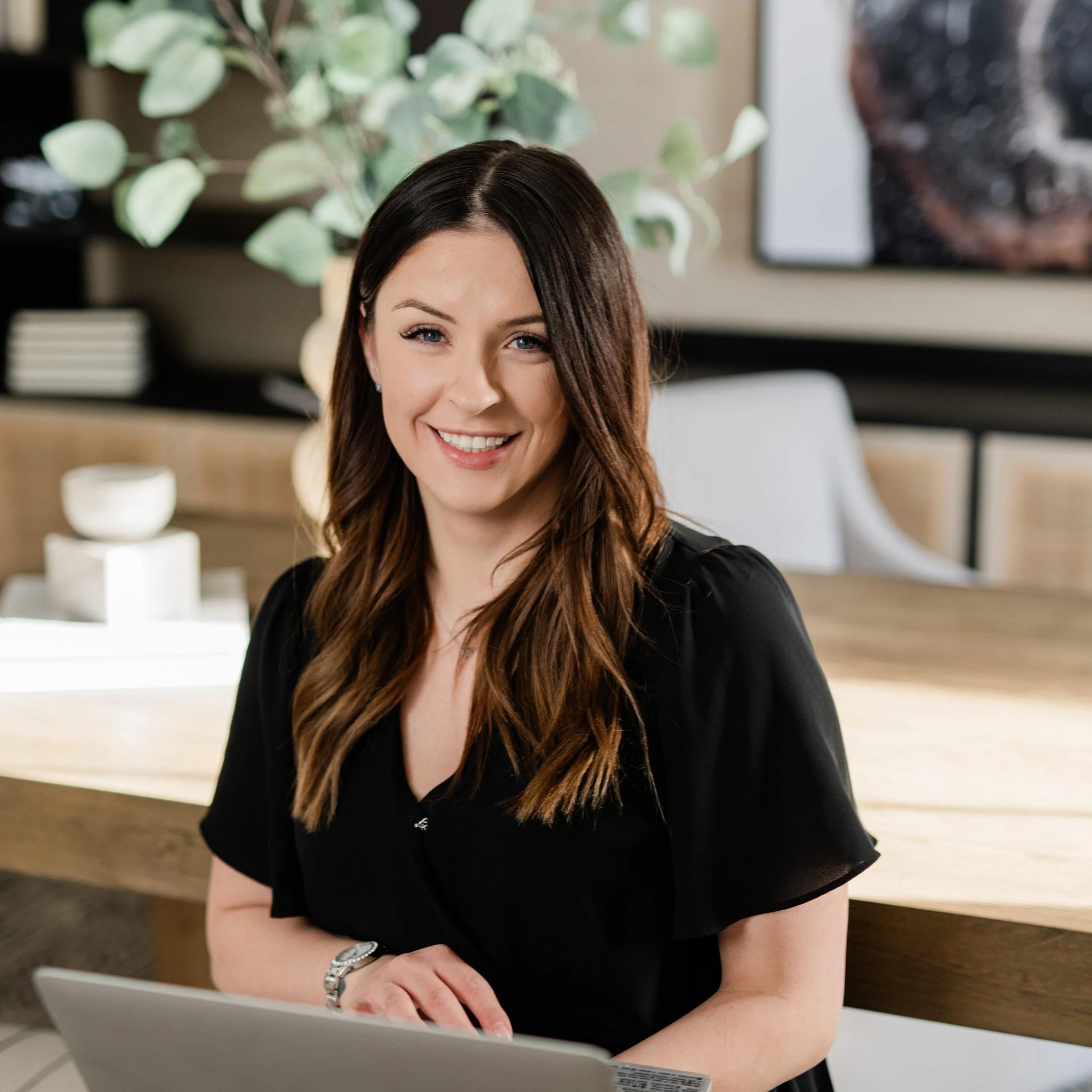 A woman with long brown hair smiling while sitting at a desk with a laptop inside an office.