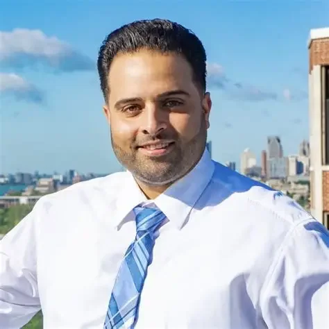 A man with dark hair and a beard smiling outdoors, wearing a white shirt and blue tie, with a city skyline and blue sky in the background.