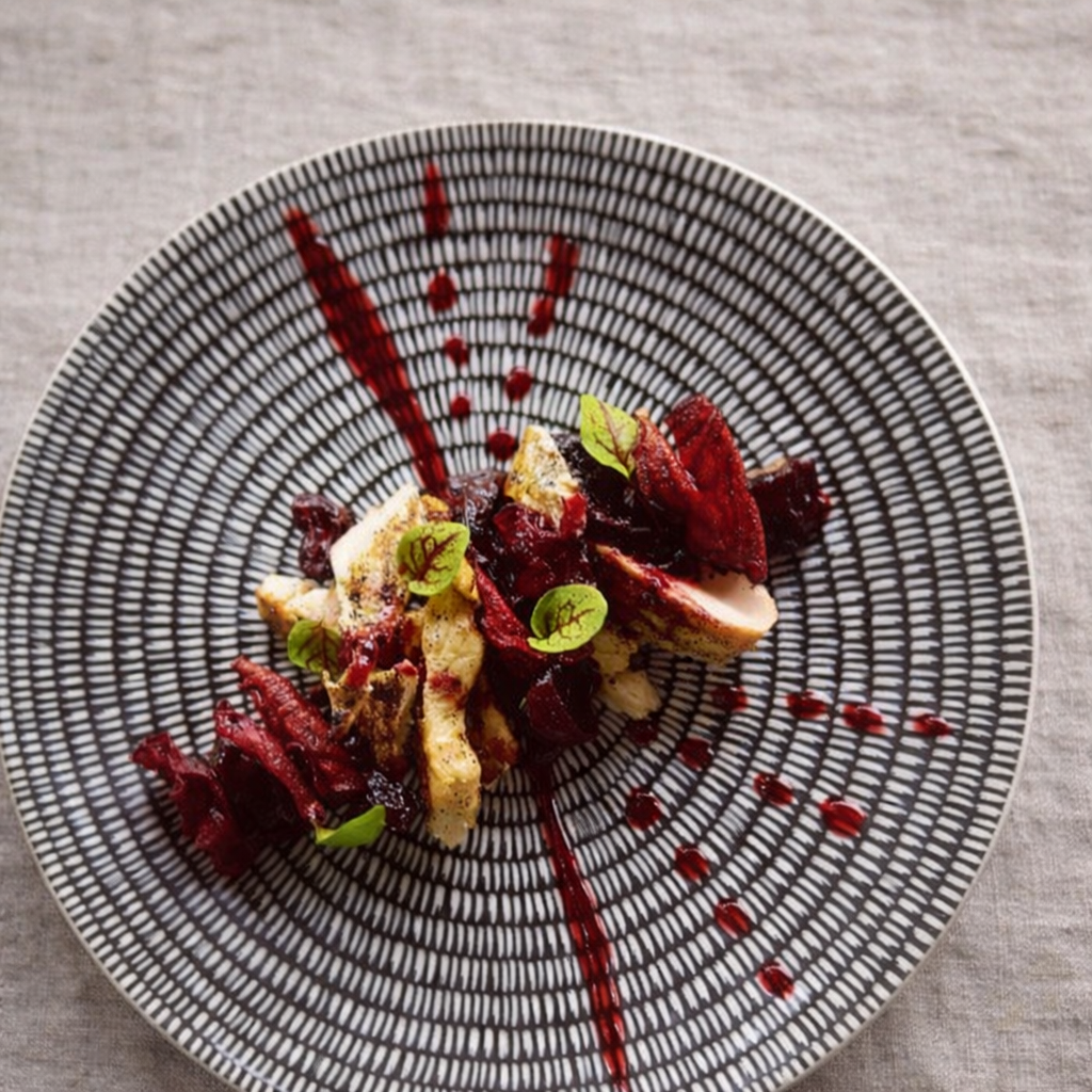A gourmet dish with smoked chicken, beetroot, micro herbs,raspberry sauce on a patterned black and white plate.
