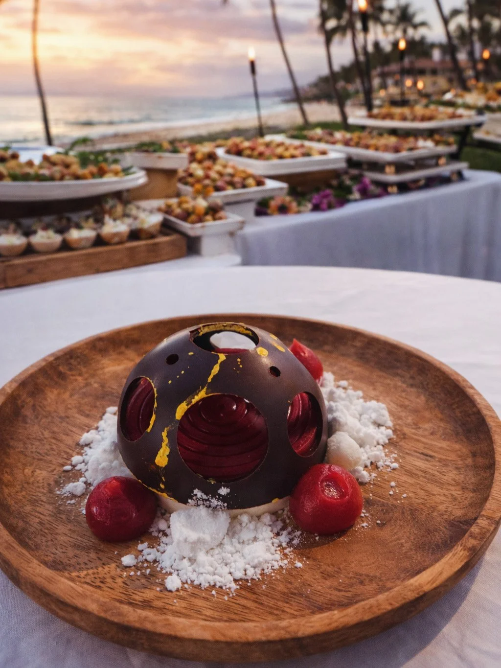 Chocolate spherical dessert on a wooden plate with red fruit and white chocolate snow powder, with a beach and sunset in the background and tables of assorted desserts.
