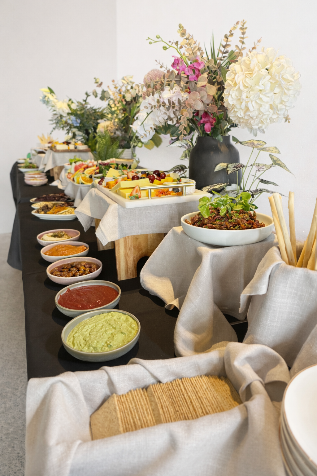 A buffet table with a floral centerpiece featuring white and pink flowers and greenery, with various dishes like cheeses, fruits, dips, and snacks arranged on it.