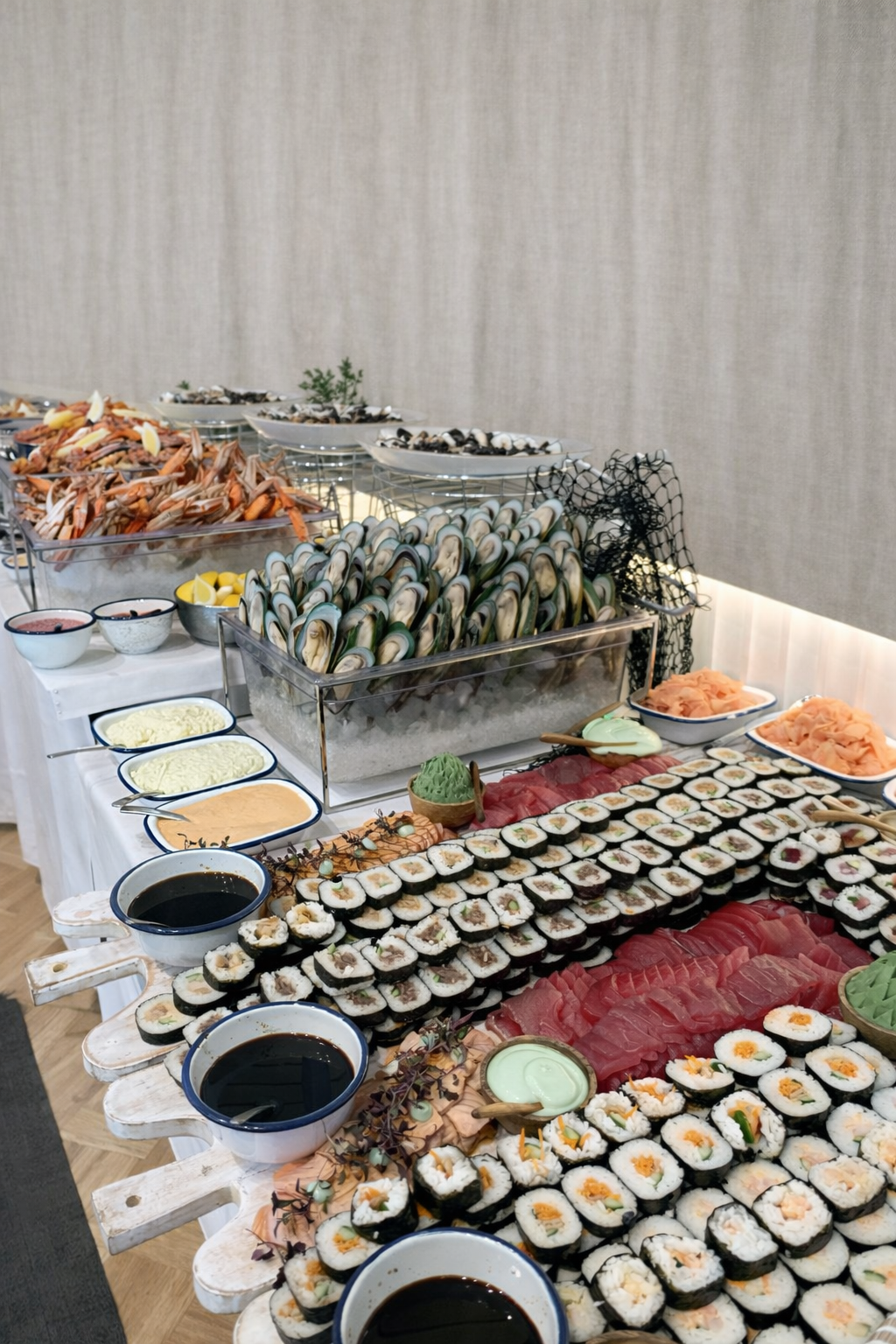 A buffet table with various sushi rolls, sashimi, oysters, prawns, crabs, mussels and condiments, and dipping sauces displayed on a white tablecloth.