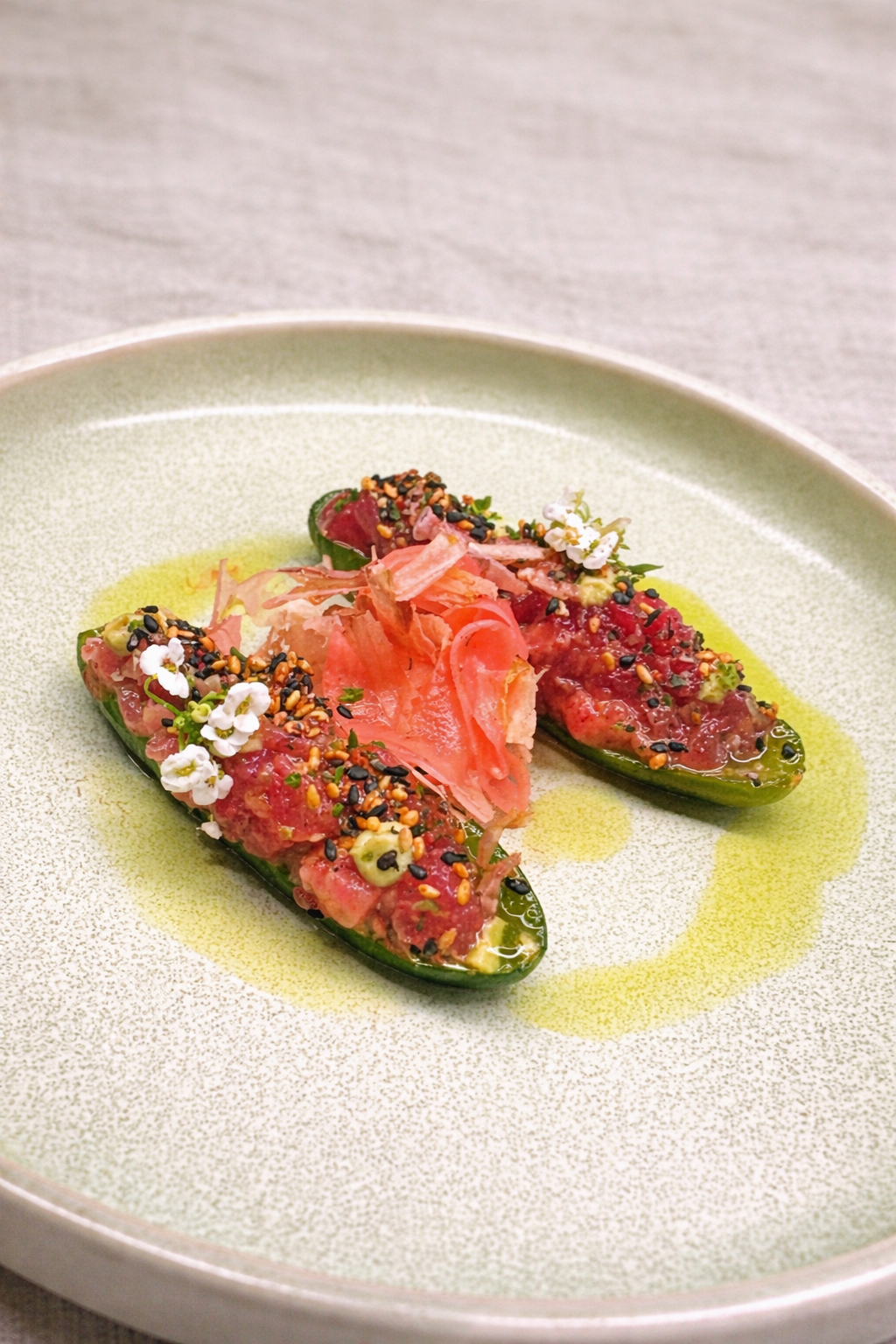 Two compressed baby cucumber filled with a mixture of tuna, topped with sesame seeds, chopped herbs, and edible flowers, served with a drizzle of oil on a green plate.