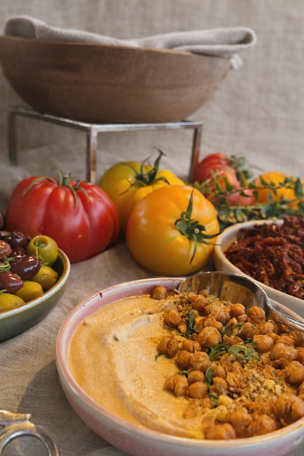 Close-up of a table filled with colourful heirloom tomatoes, bowls of sundried tomatoes, chickpea dip, marinated olives, with a wooden bowl and cloth napkin in the background.