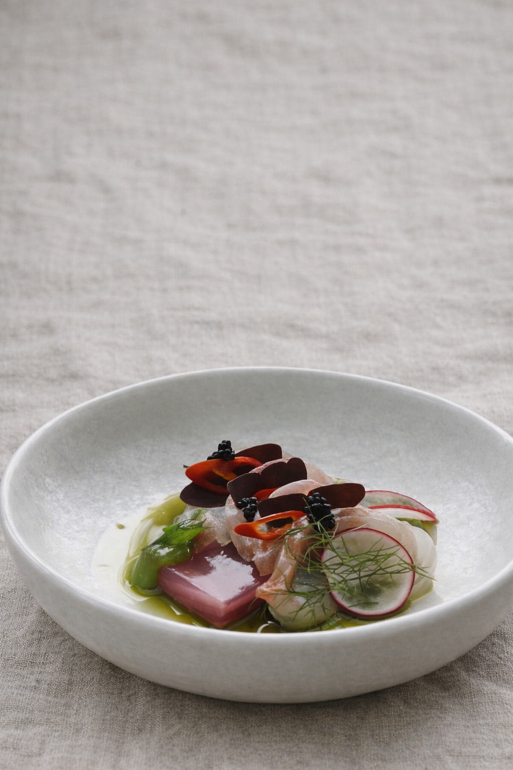 A white ceramic bowl containing sliced radishes, baby snapper, green herb oil, and raw fish, garnished with microgreens, fished with chilli oil, on a textured beige surface.