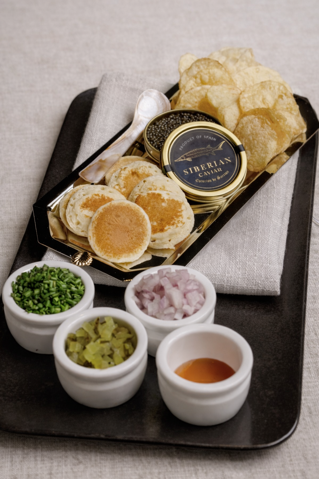 A tray with blini topped with caviar, a tin of Siberian caviar, potato chips, and small bowls of chopped onions, chives, pickles, and horseradish. Small spoon included.