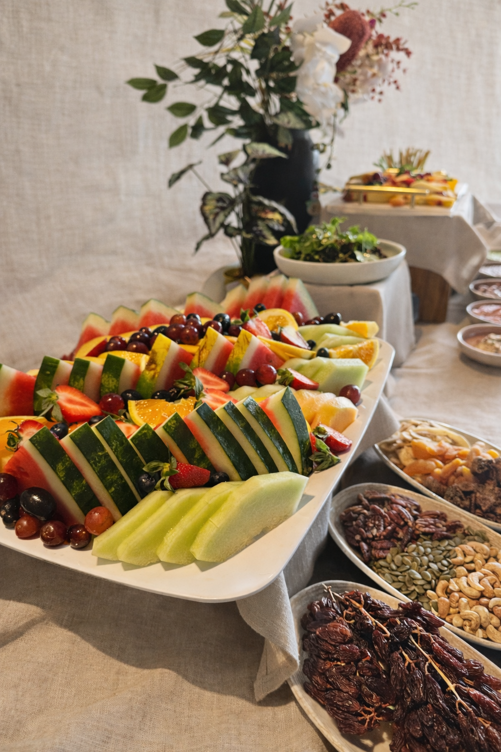 A table with various fresh fruits, including watermelon, rockmelon, honeydew, strawberries, grapes, and oranges, arranged on a large platter. There are also bowls of nuts, dried dates, and other snacks, along with a vase of flowers in the background.