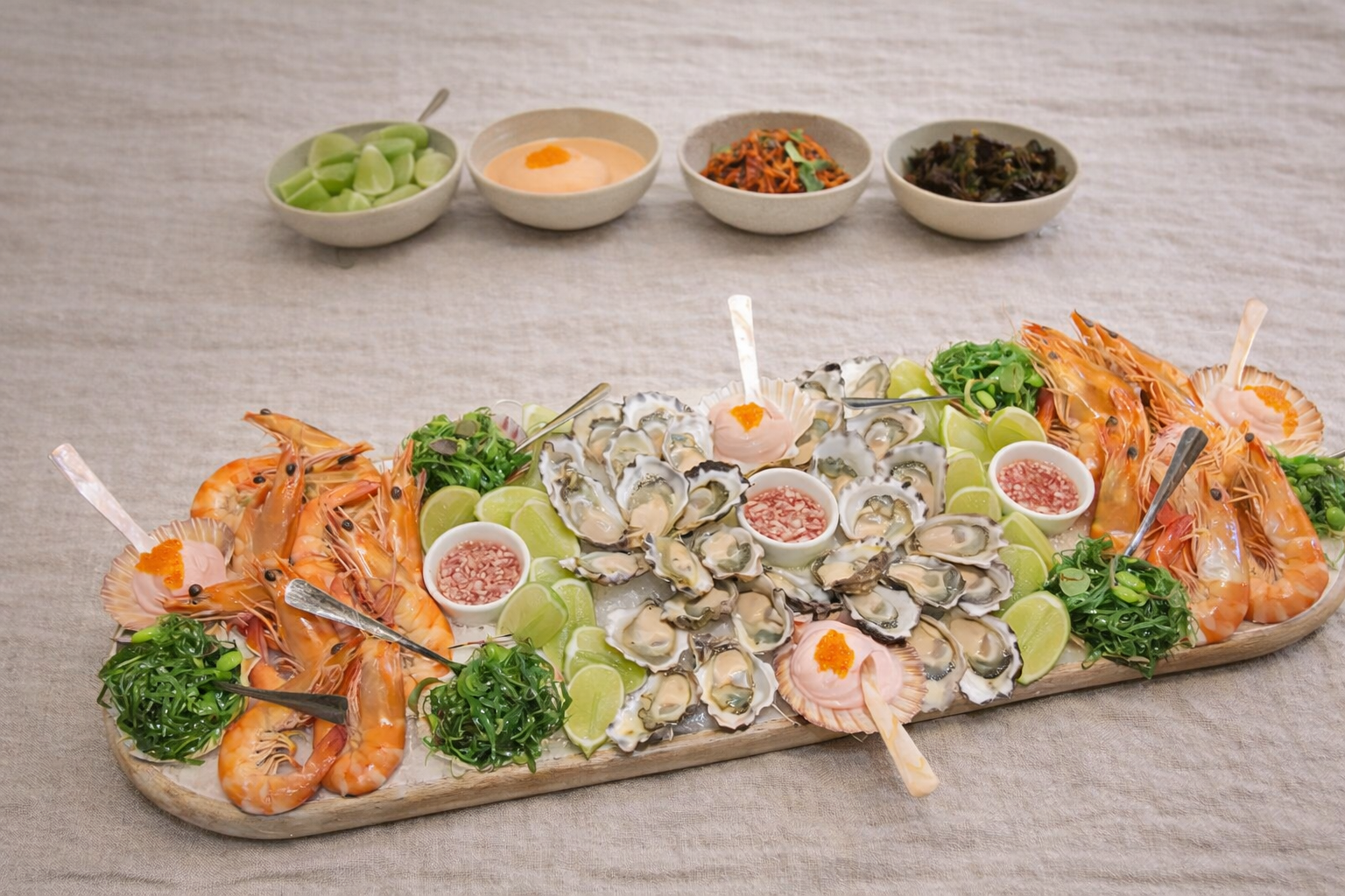Seafood platter featuring oysters, prawns, lime wedges, and dipping sauces, with side bowls of sauce and condiments in the background.