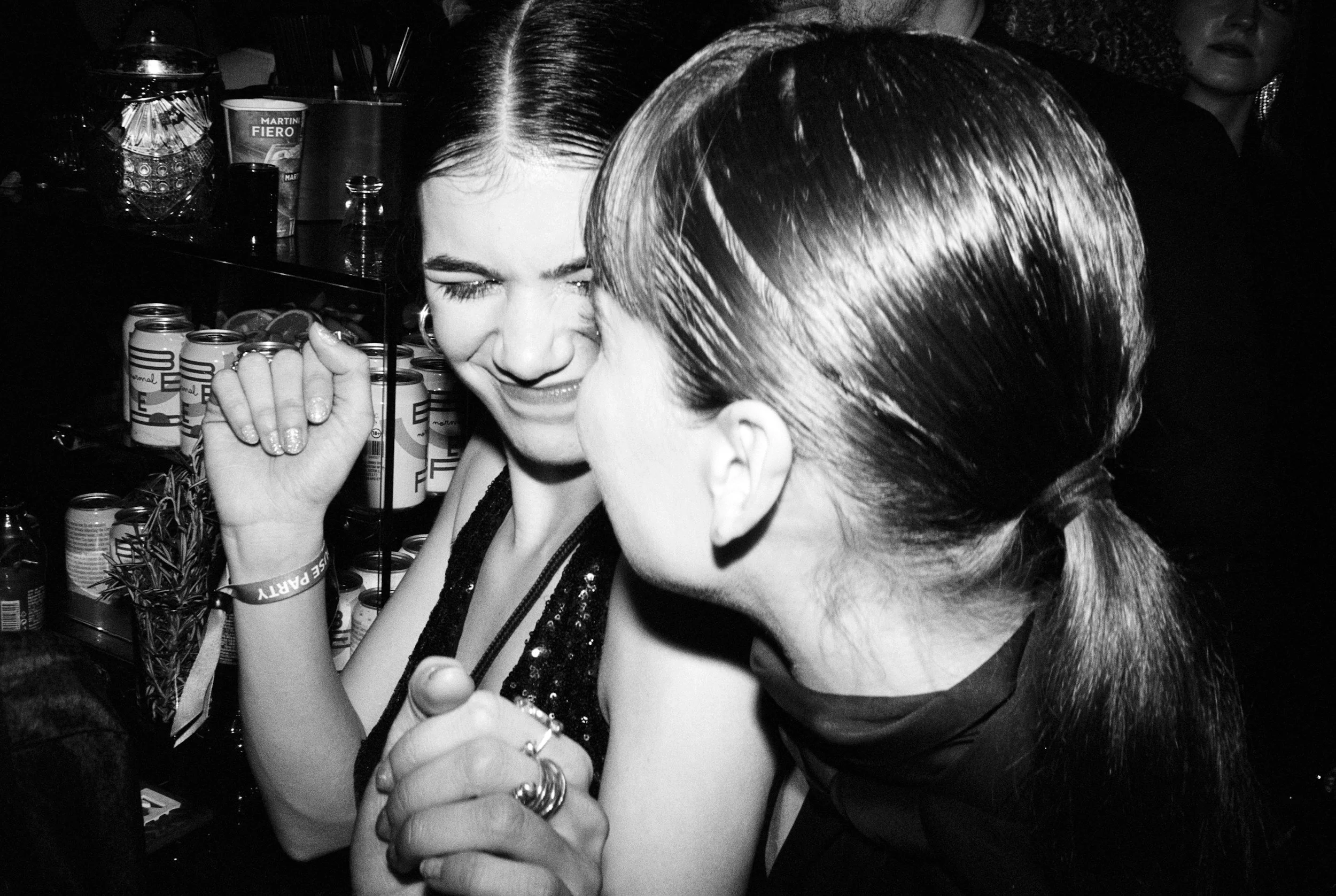 Two women at a social gathering, laughing and leaning close to each other with drinks in the background.