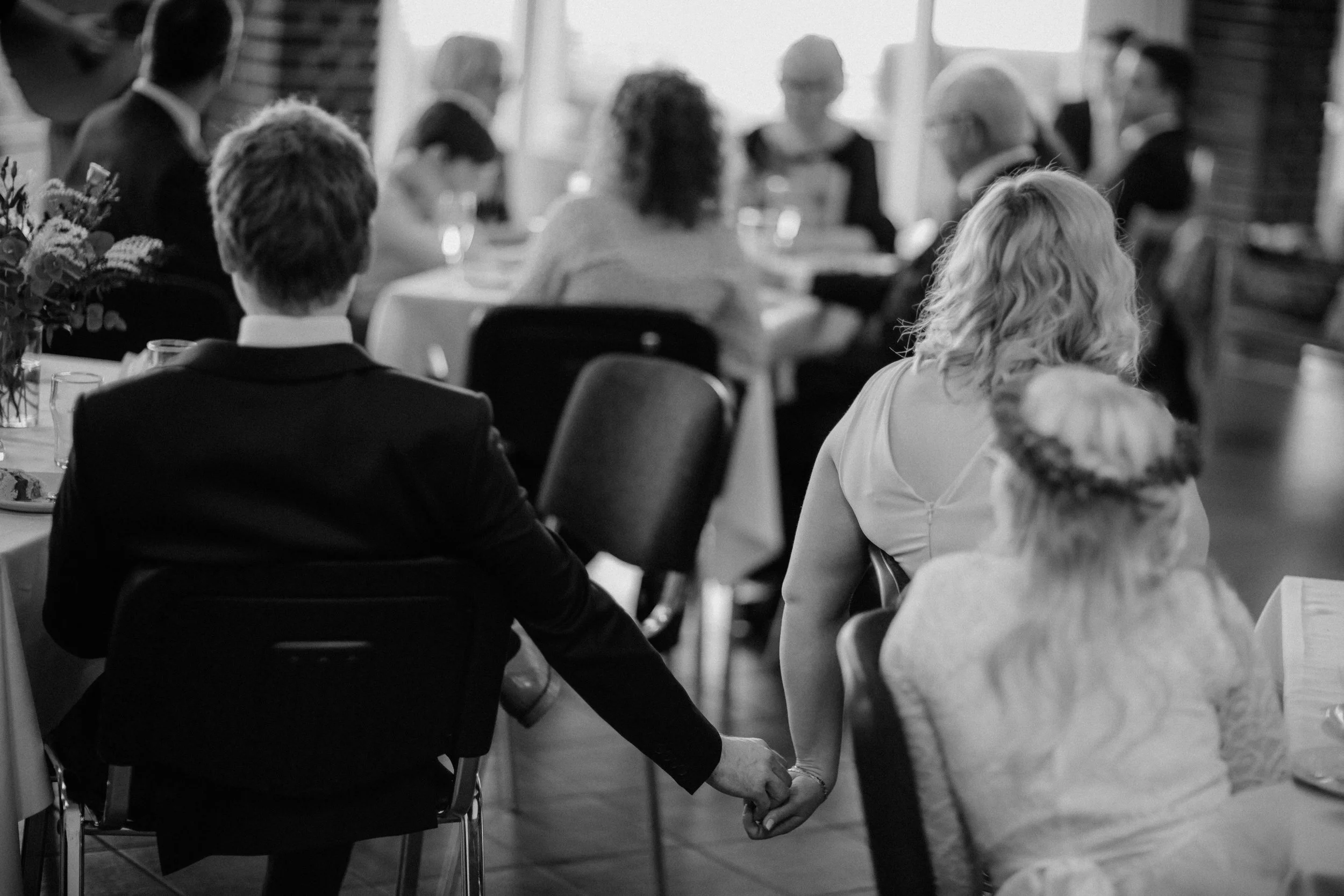 A couple holding hands at a wedding reception, sitting among guests at a decorated table.