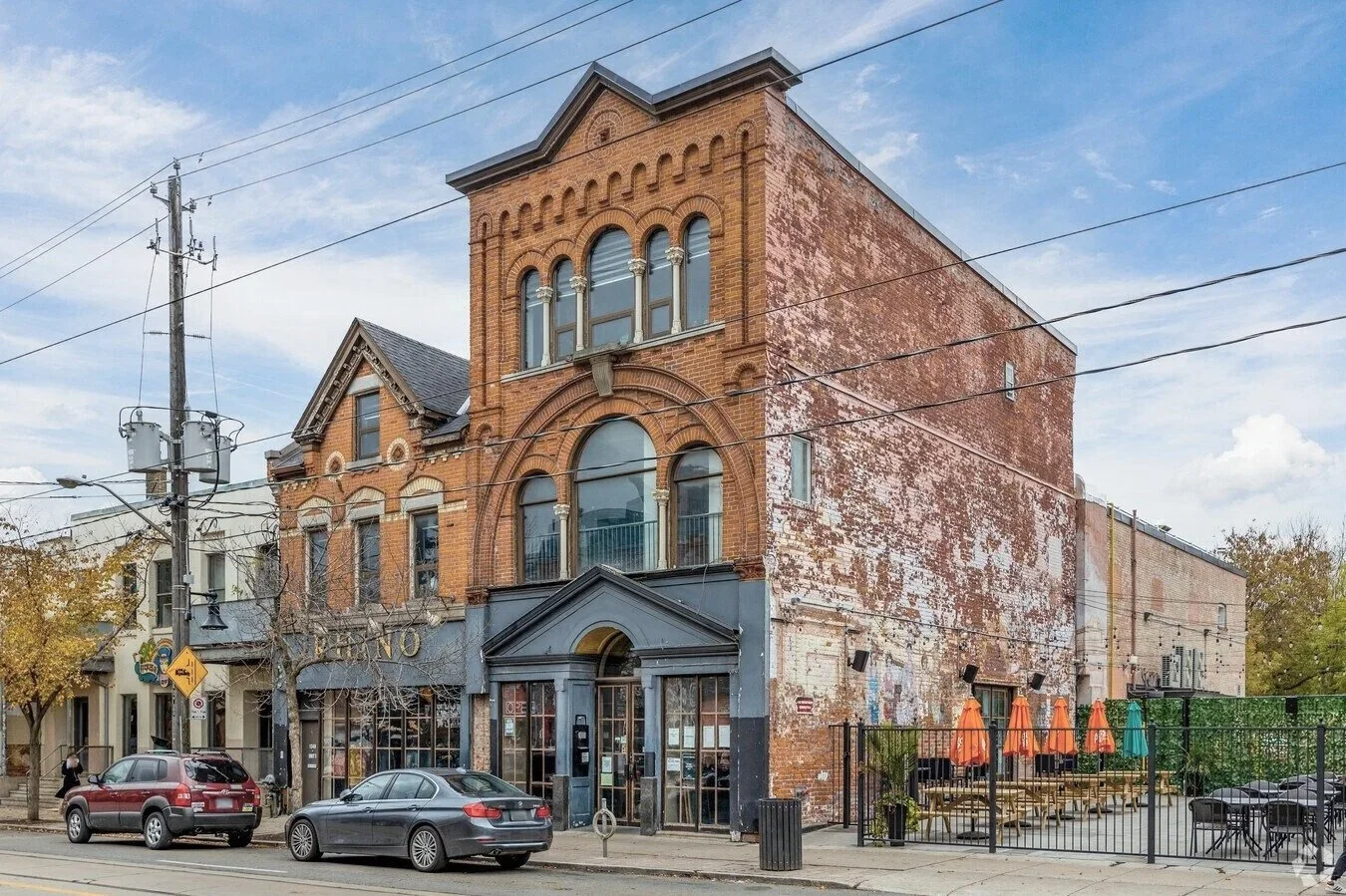 Older brick building with arched windows and decorative architectural details, with a neighboring building and cars parked outside, and an outdoor seating area with orange and blue umbrellas.