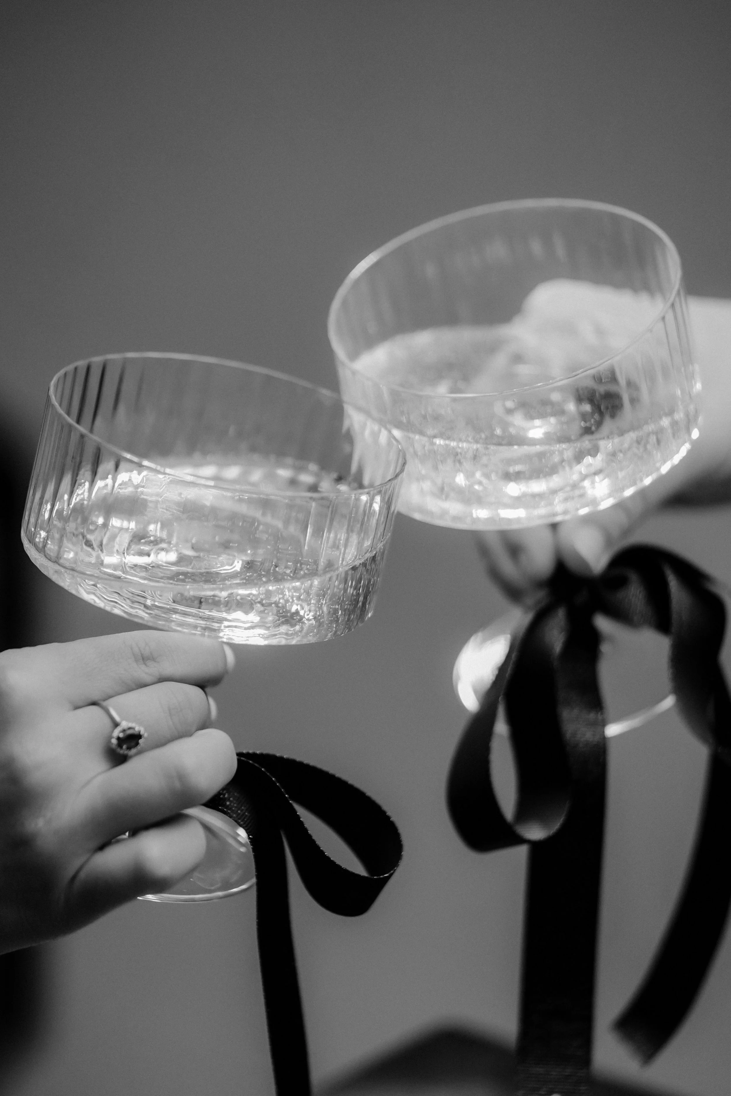 Two champagne glasses filled with a clear liquid, held by hands with ribbons tied to their stems, in a celebratory toast.