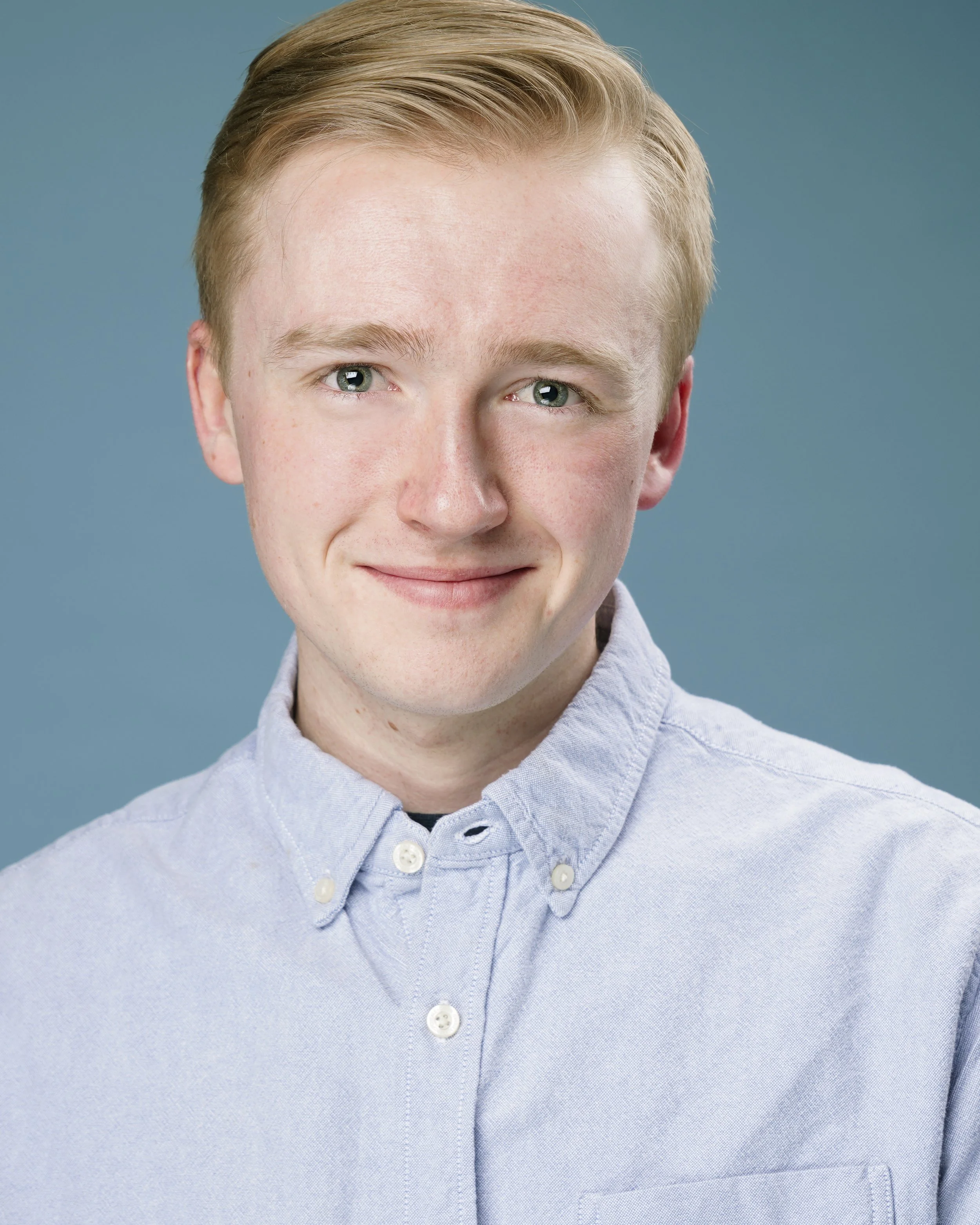 A young man with light skin, short blond hair, and blue eyes, smiling while wearing a light blue button-up shirt against a solid blue background.