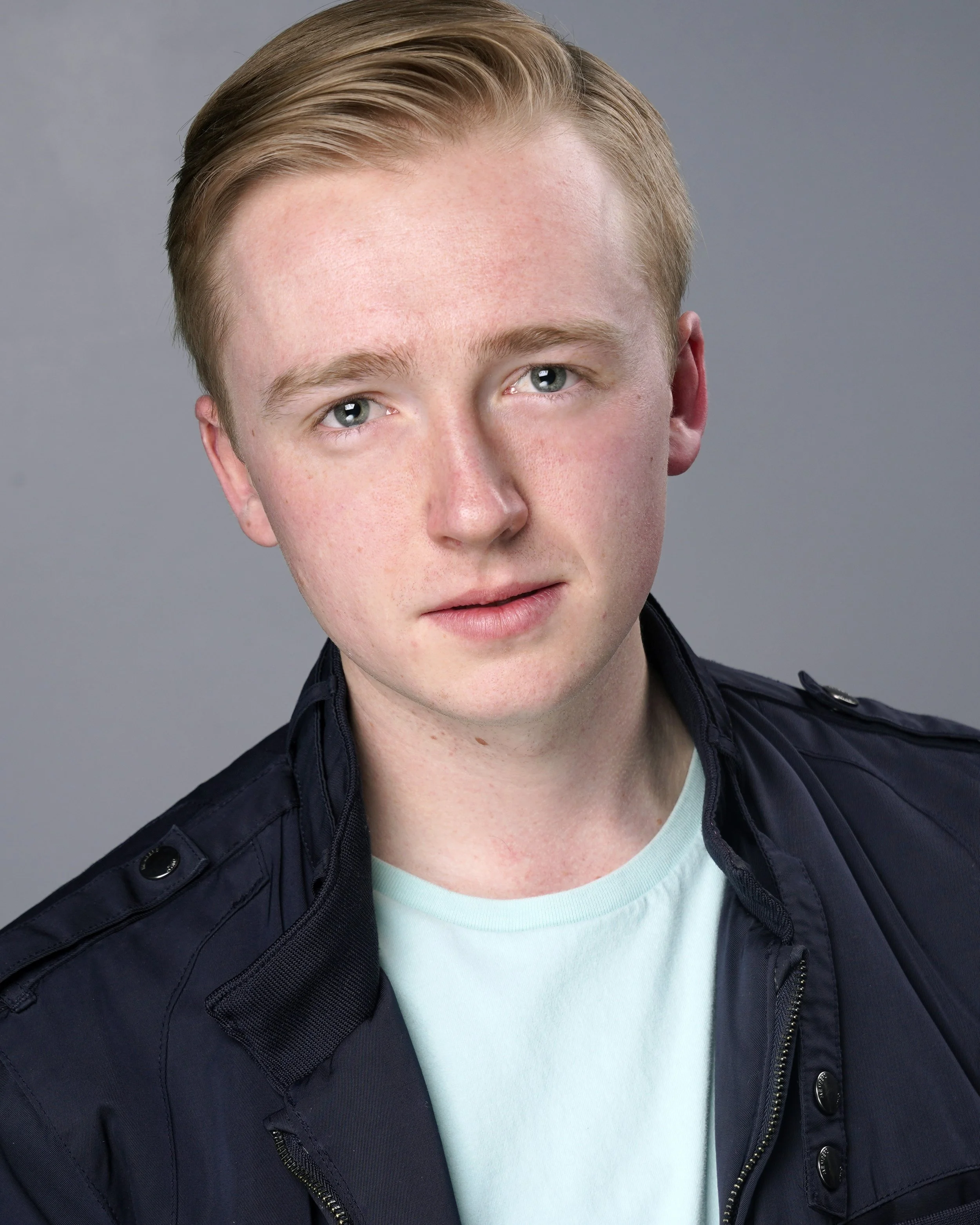 A young man with fair skin and blond hair styled to the side, wearing a light blue t-shirt and a dark navy jacket, against a plain gray background.