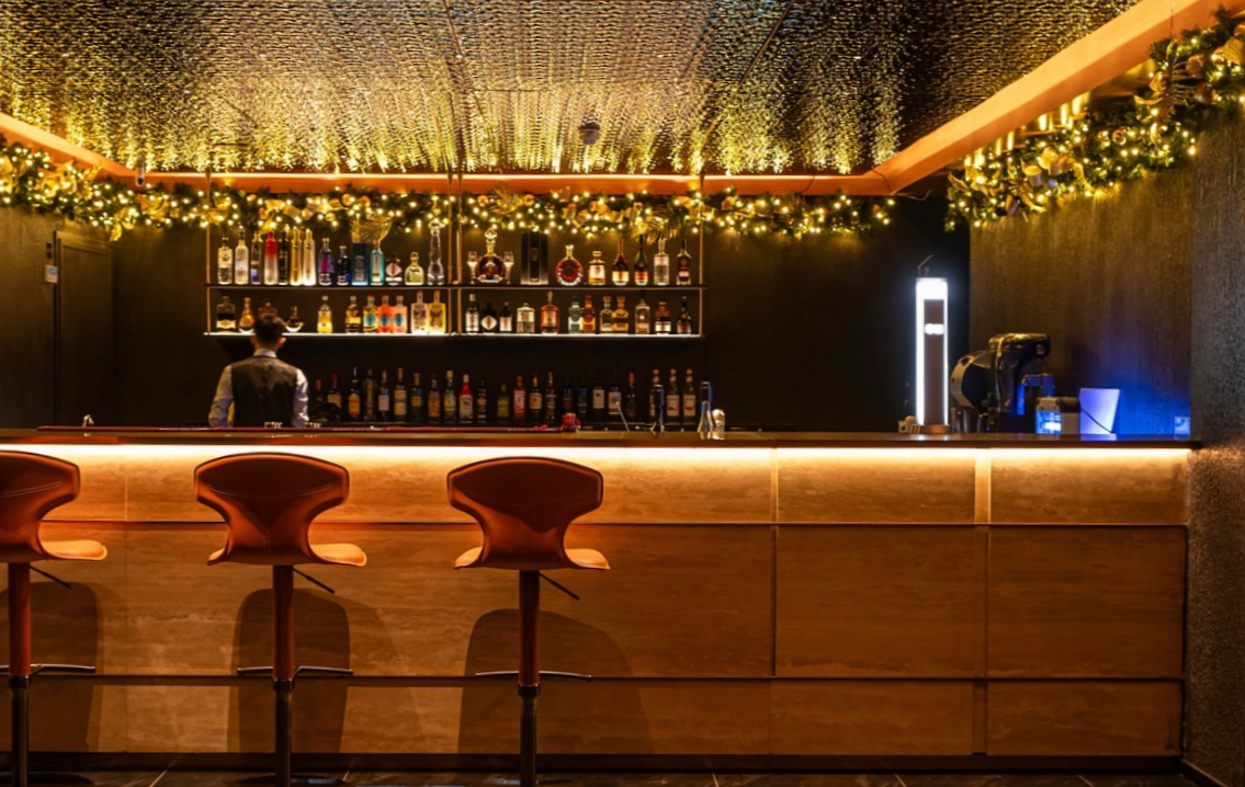 An elegant bar with a wooden counter, illuminated with warm lighting, decorated with holiday garlands with lights, and stocked with various bottles of alcohol on shelves behind the bar. Three wooden bar stools are placed in front of the counter, and a bartender is working behind the bar. The ceiling is decorated with a textured, shiny gold pattern.