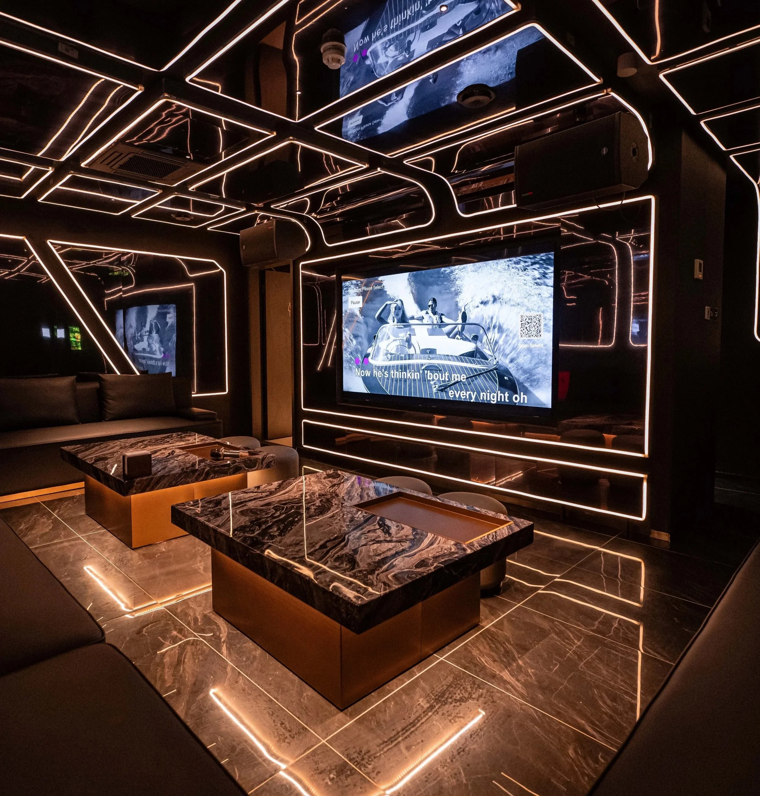 A private karaoke room illuminated by geometric light strips, furnished with black leather sofas and marble-topped coffee tables. The room features reflective marble flooring and a cosy, stylish atmosphere.