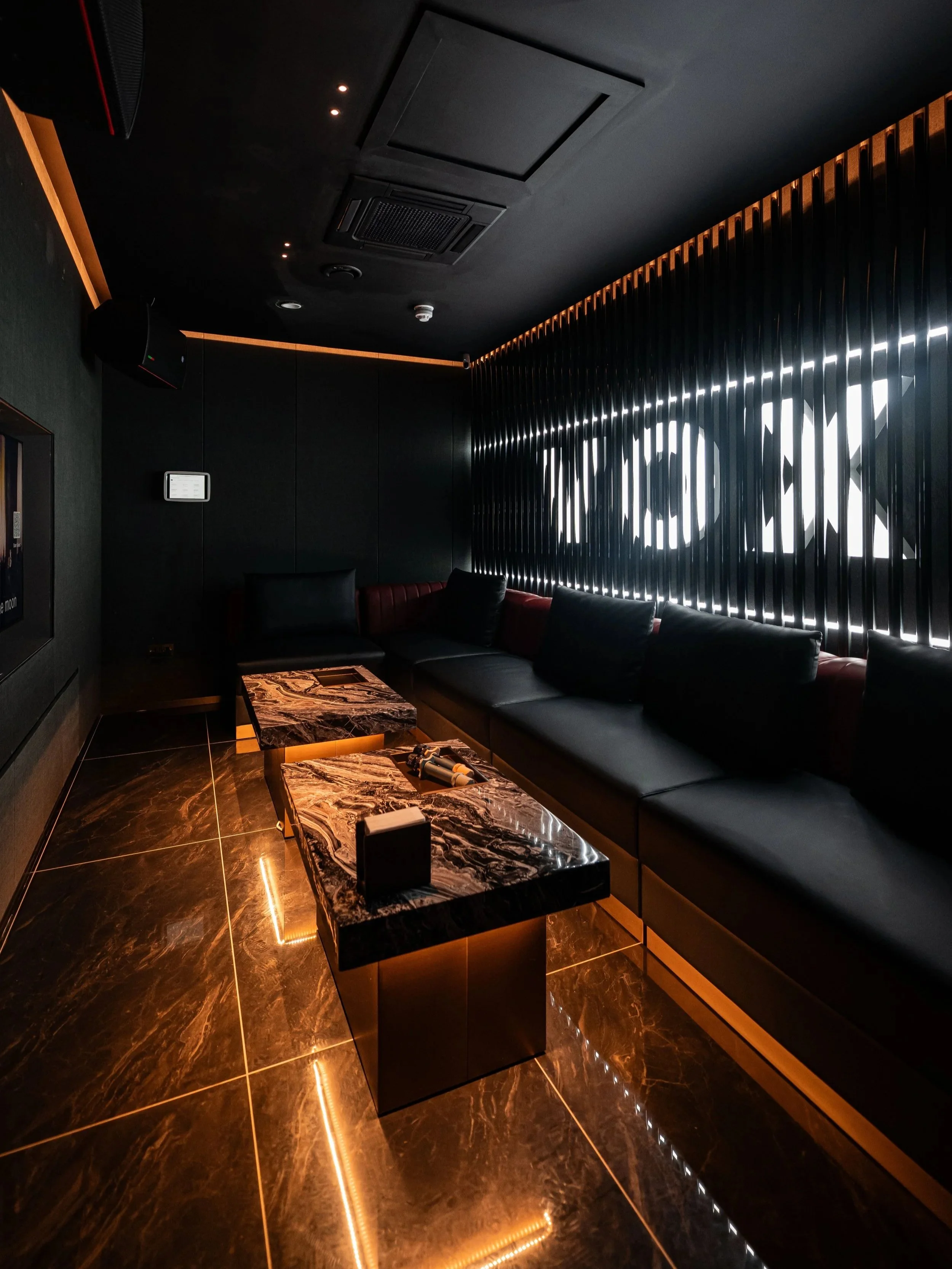 A private karaoke room with plush leather seating, marble tables, and illuminated black slatted wall with the VOX Karaoke Cocktail Bar logo.