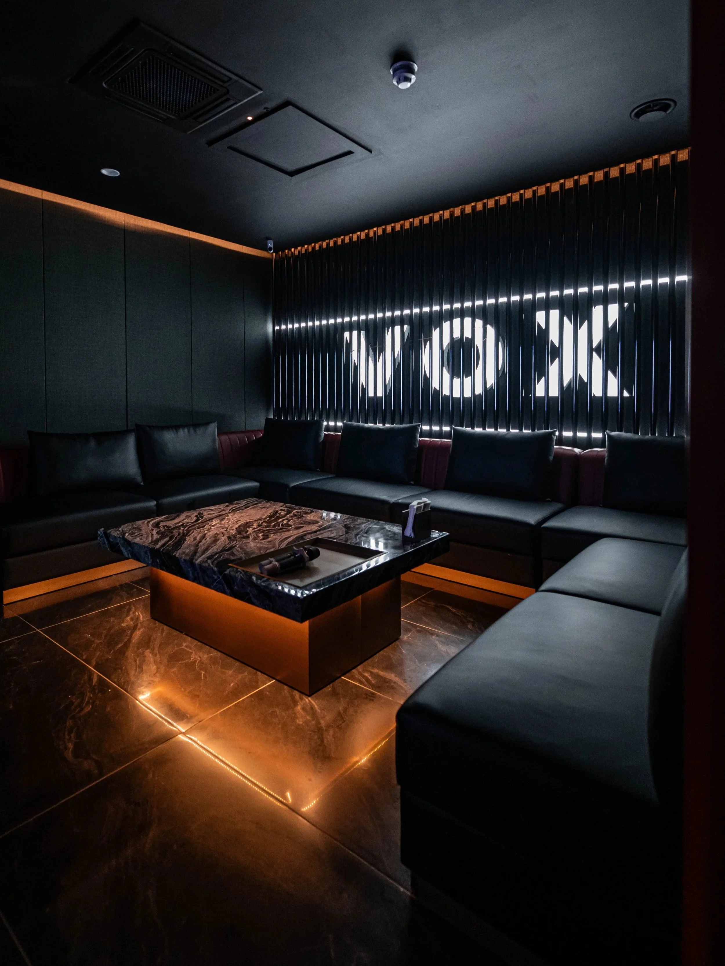 A private karaoke room fitted with plush leather sofas and a black marble coffee table. The back wall features a vertical slat design with an illuminated 'VOX' logo behind it.