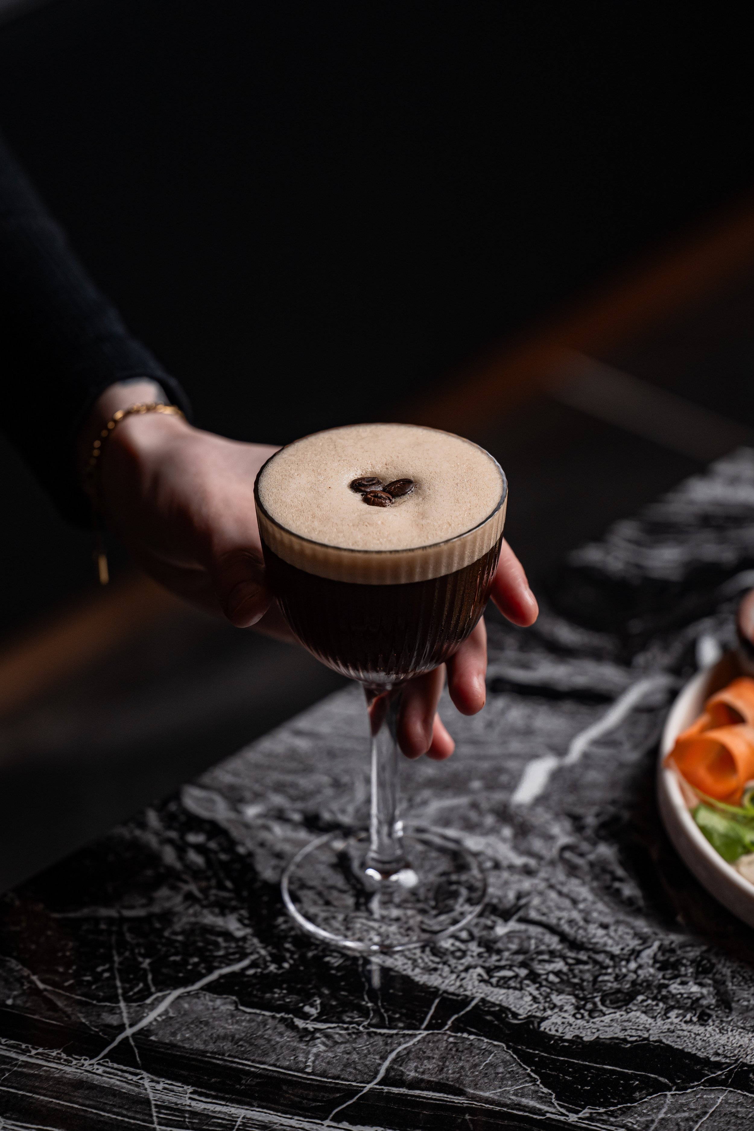A person holding an Espresso Martini, on a marble table next to a plate of food.