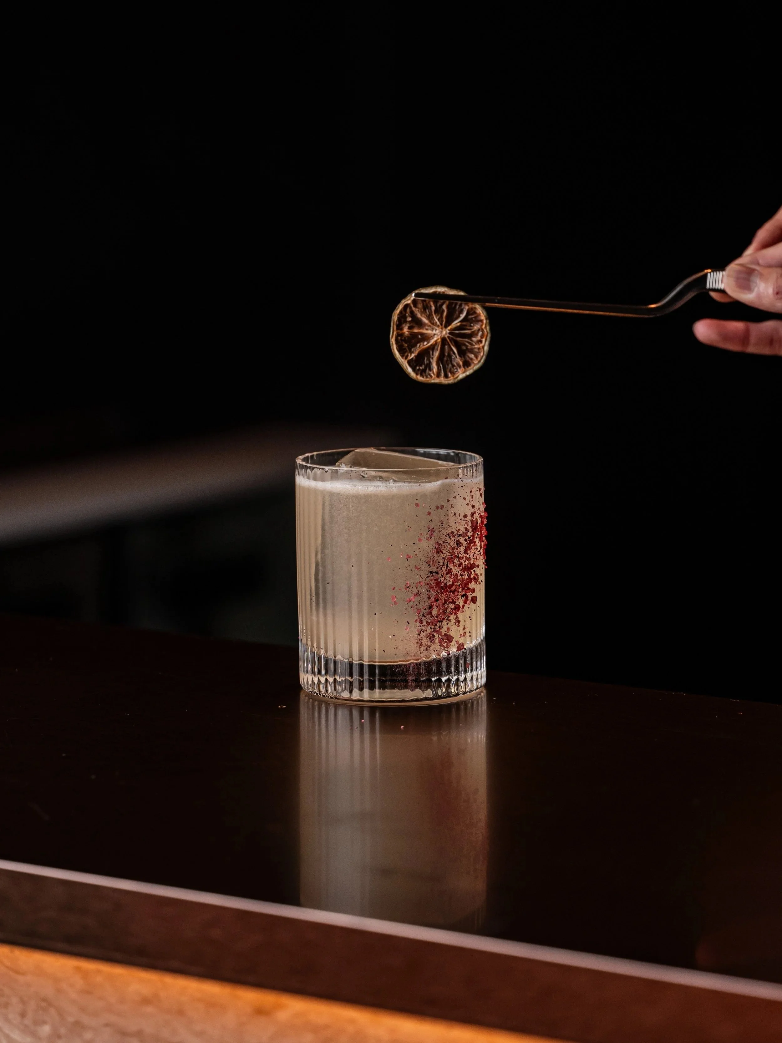 A bartender is garnishing our Belle Margarita with a dried lime slice using tongs. The cocktail is in a hand-blown ribbed glass with hibiscus salt sprinkled on the side.