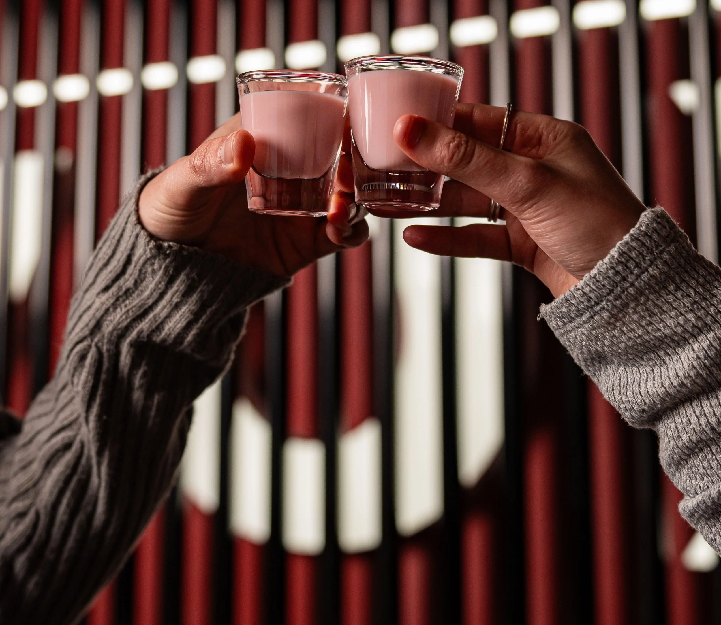 Two hands holding shot glasses filled with a pink beverage against a blurred background of alternating red and white vertical slats.
