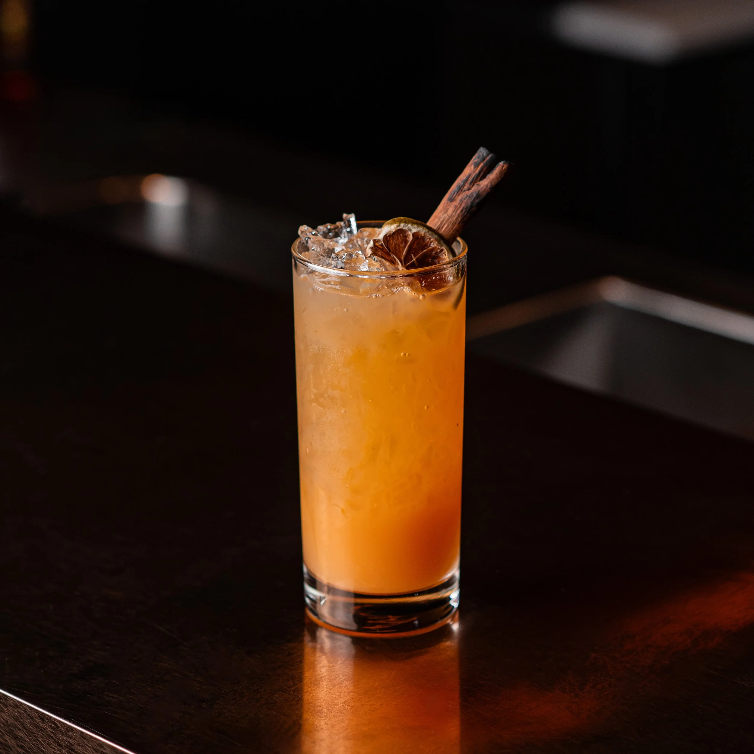 A Tropical Sunset cocktail, topped with crushed ice, garnished with a slice of dried lime and a cinnamon stick.