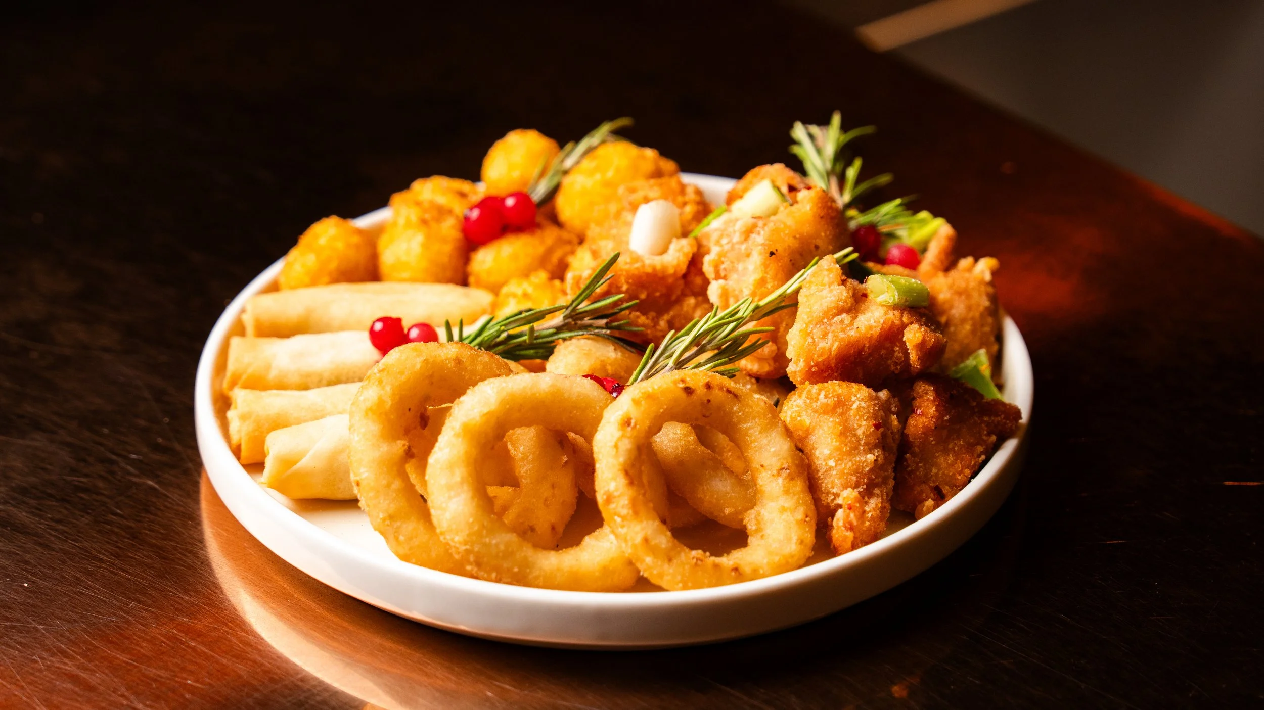 A white bowl filled with fried onion rings, mozzarella sticks, fried chicken pieces, garnished with sprigs of rosemary and red berries, on a dark wooden surface.