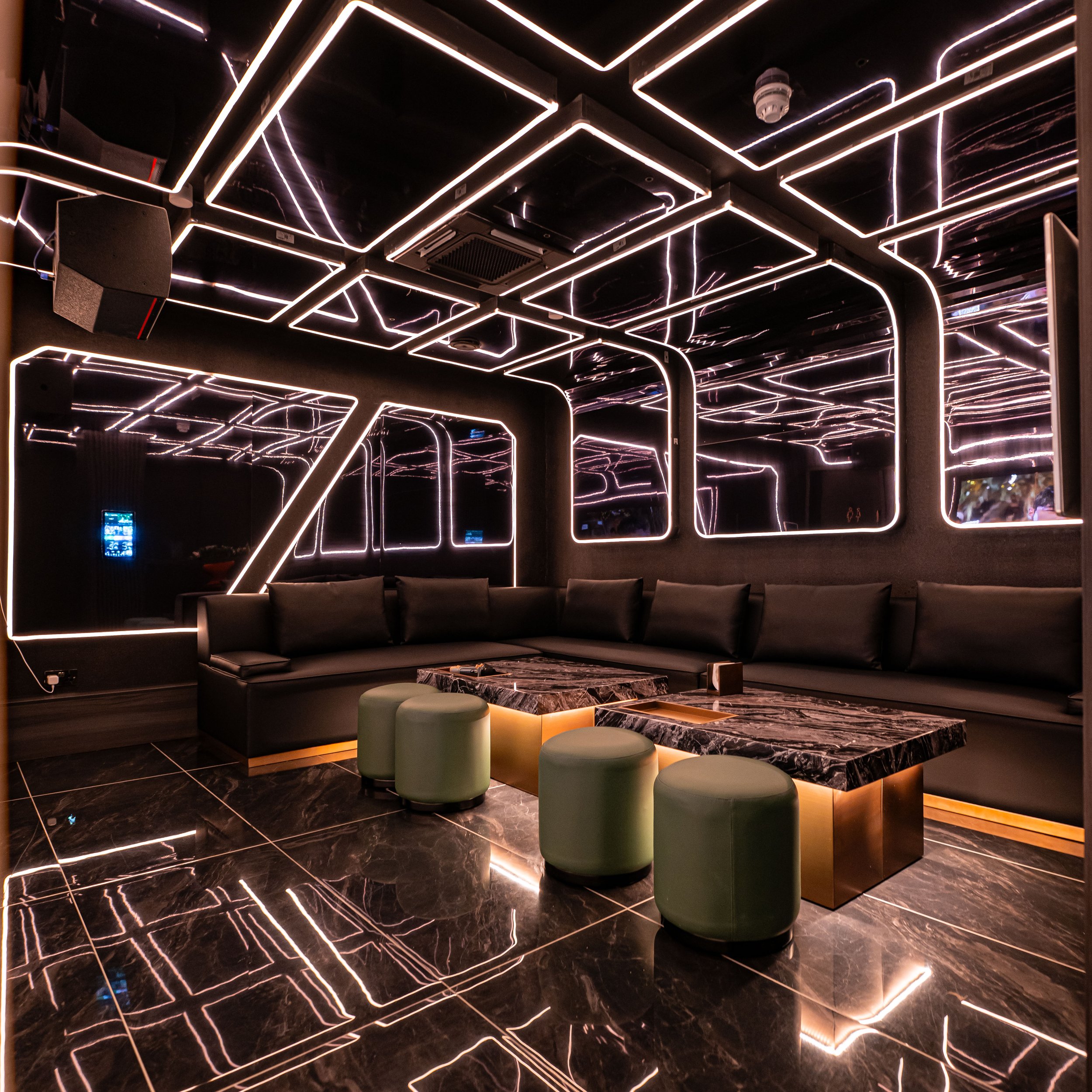 Interior of a modern lounge or club with black marble flooring, black walls, and black ceiling adorned with white neon line lights arranged in geometric patterns. There is a black leather sectional couch along the wall, a marble coffee table, and several round light green ottomans in the foreground. Mirrors reflect the neon lighting, creating a futuristic ambiance.