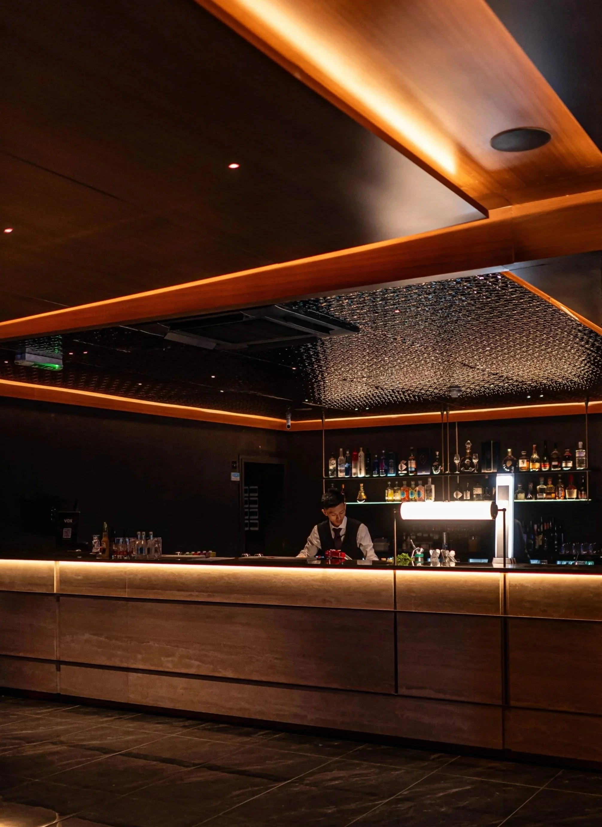 A bartender preparing drinks behind a bar counter with cosy, warm lighting in VOX Karaoke Cocktail Bar.
