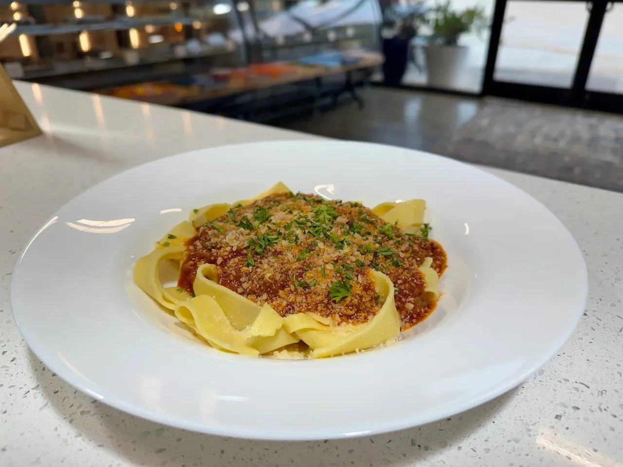 A white plate of cooked fettuccine pasta topped with marinara sauce, grated cheese, and chopped parsley, on a white countertop near a window.