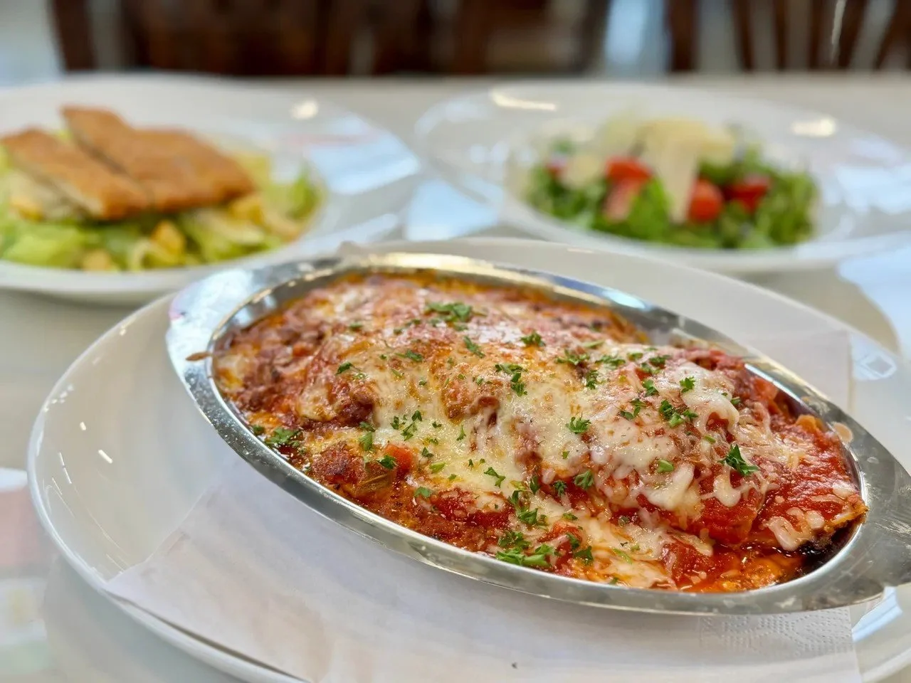 Baked lasagna with melted cheese and parsley in a metal dish, served on a white plate, with salads in the background.