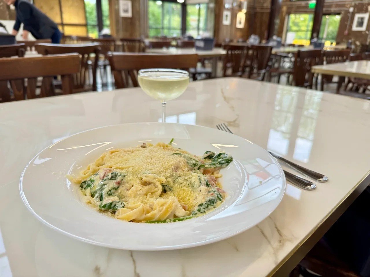 A plate of pasta topped with grated cheese, served with a glass of white wine on a white table inside a restaurant with wooden chairs and large windows.