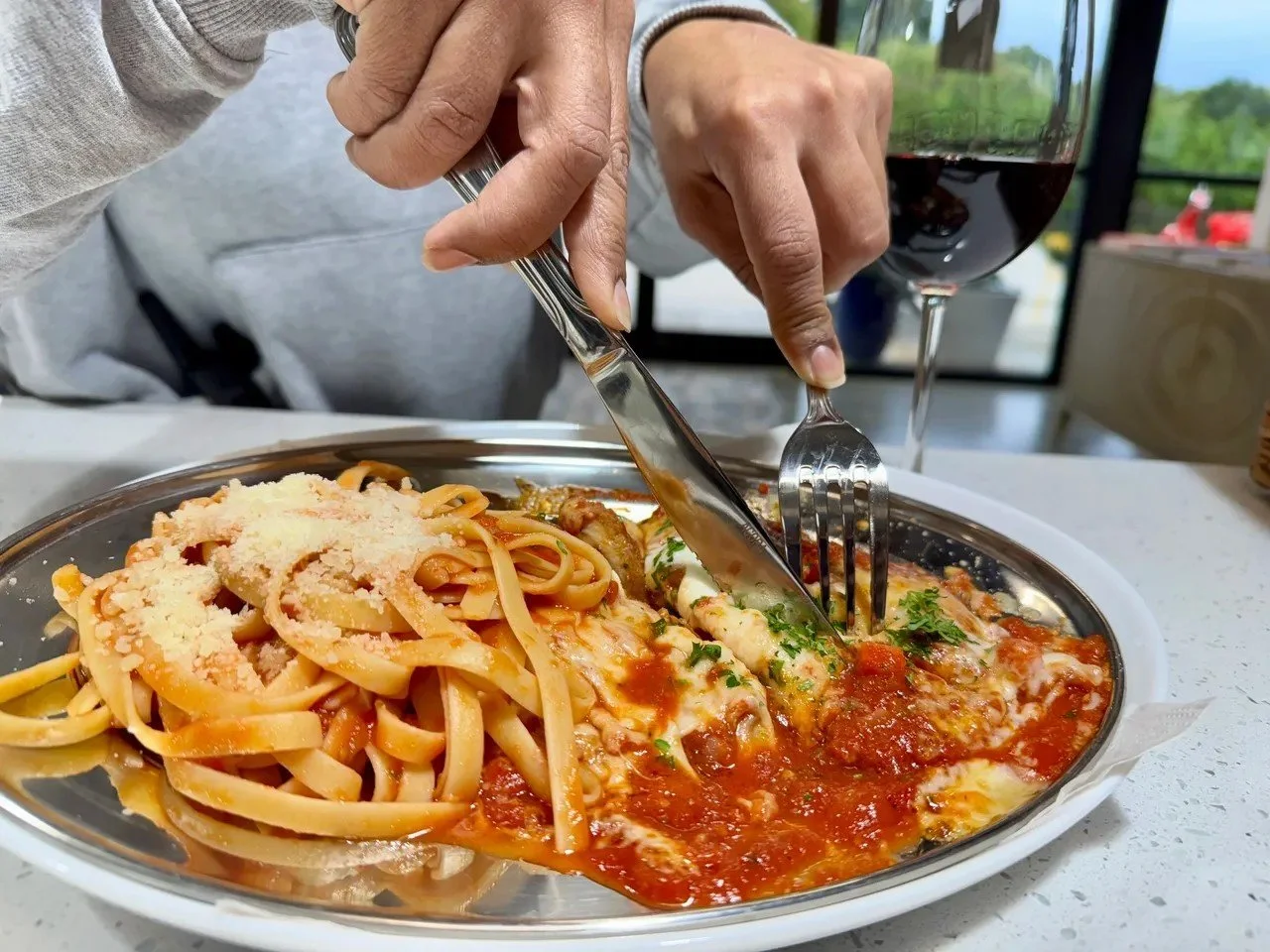 Person cutting into a piece of chicken parmesan with spaghetti on a dinner plate, alongside a glass of red wine.