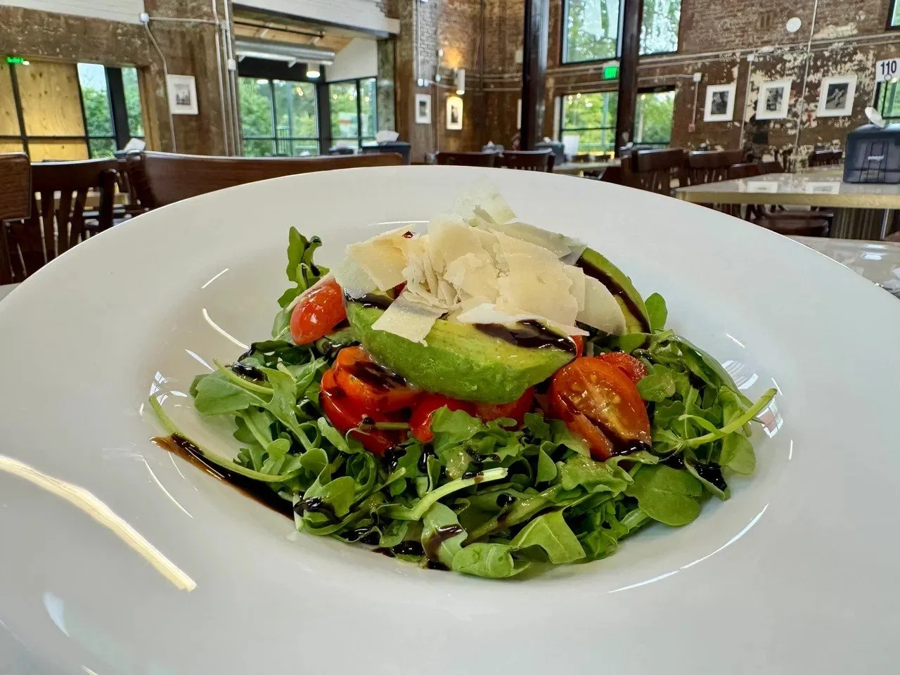 A plate of salad with arugula, cherry tomatoes, avocado, shaved Parmesan cheese, and balsamic glaze in a restaurant setting.