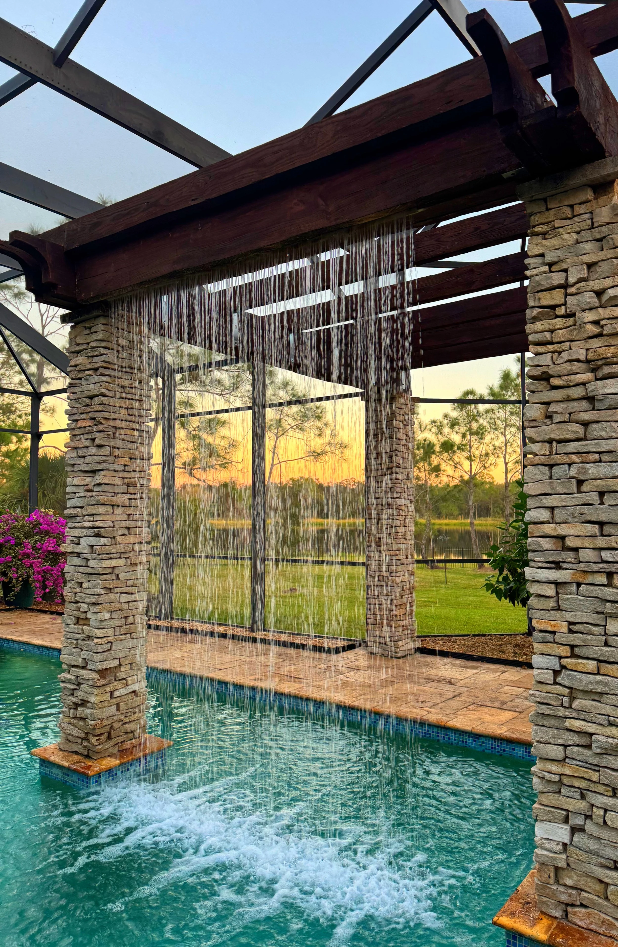 Swimming pool with a waterfall feature under a pergola, outdoors during sunset, with trees and a grassy field in the background.